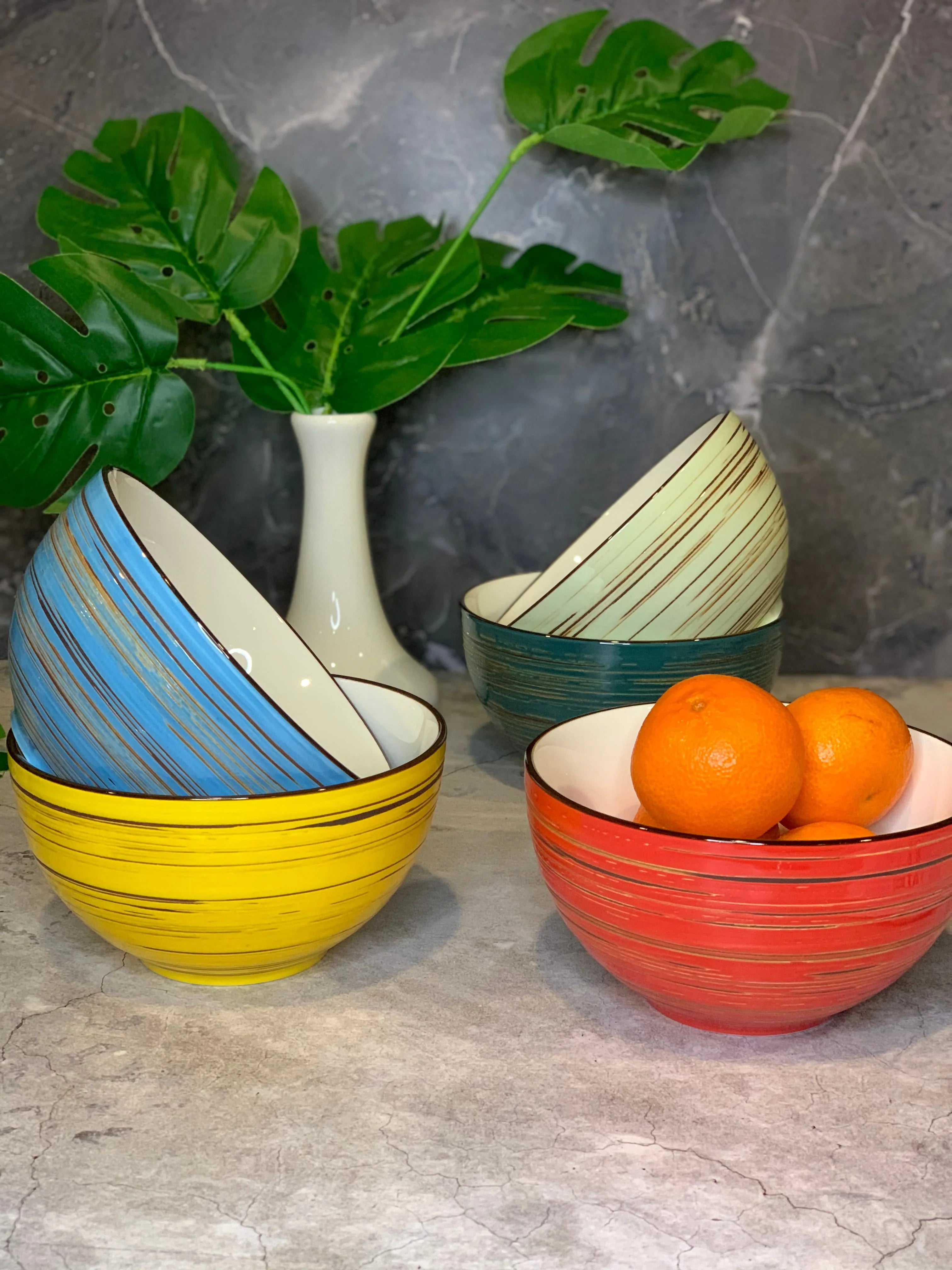 Colorful striped ceramic bowls on marble, with oranges and a white vase holding green leaves