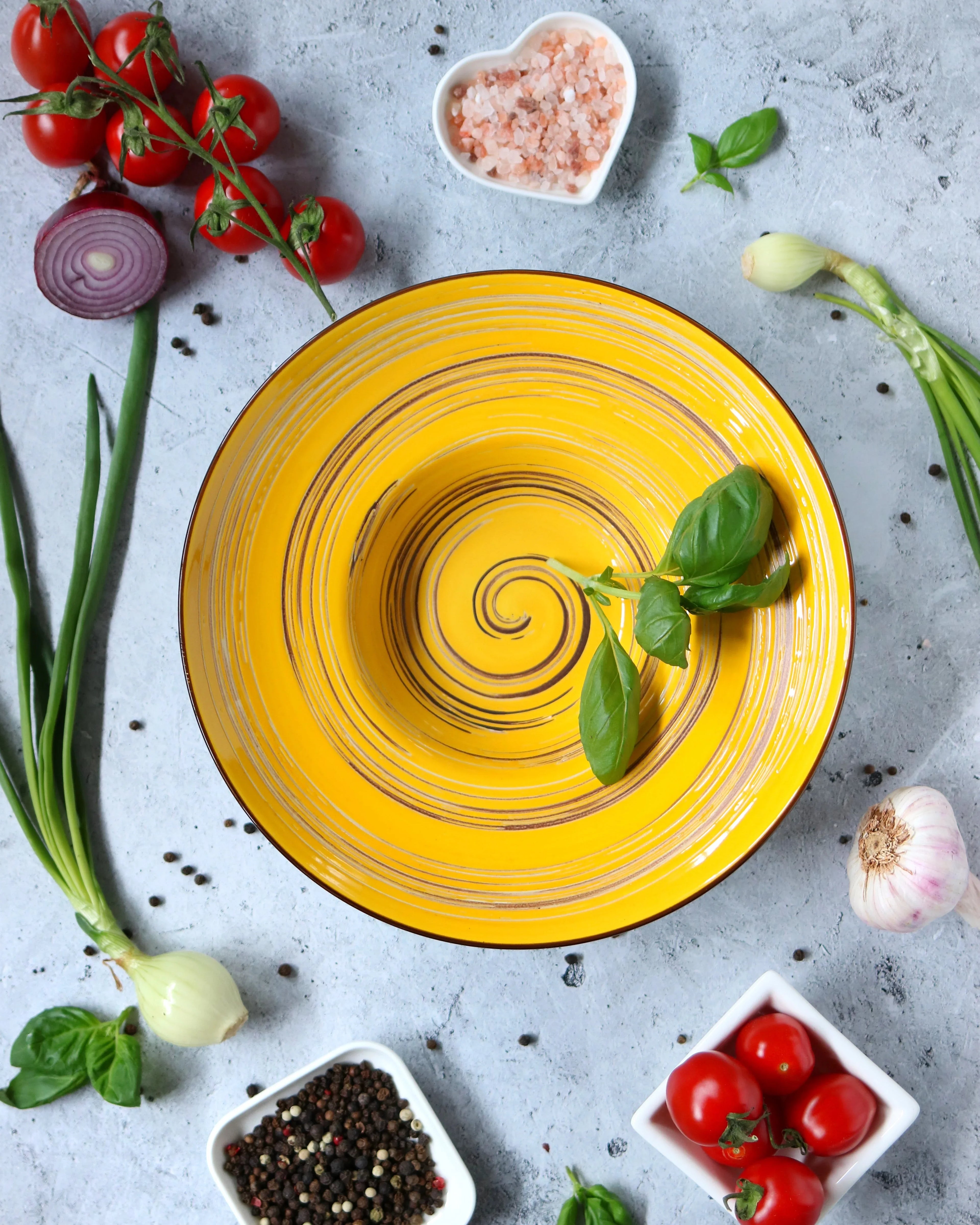 Yellow spiral artist-designed bowl with basil, surrounded by tomatoes, herbs, and seasoning.