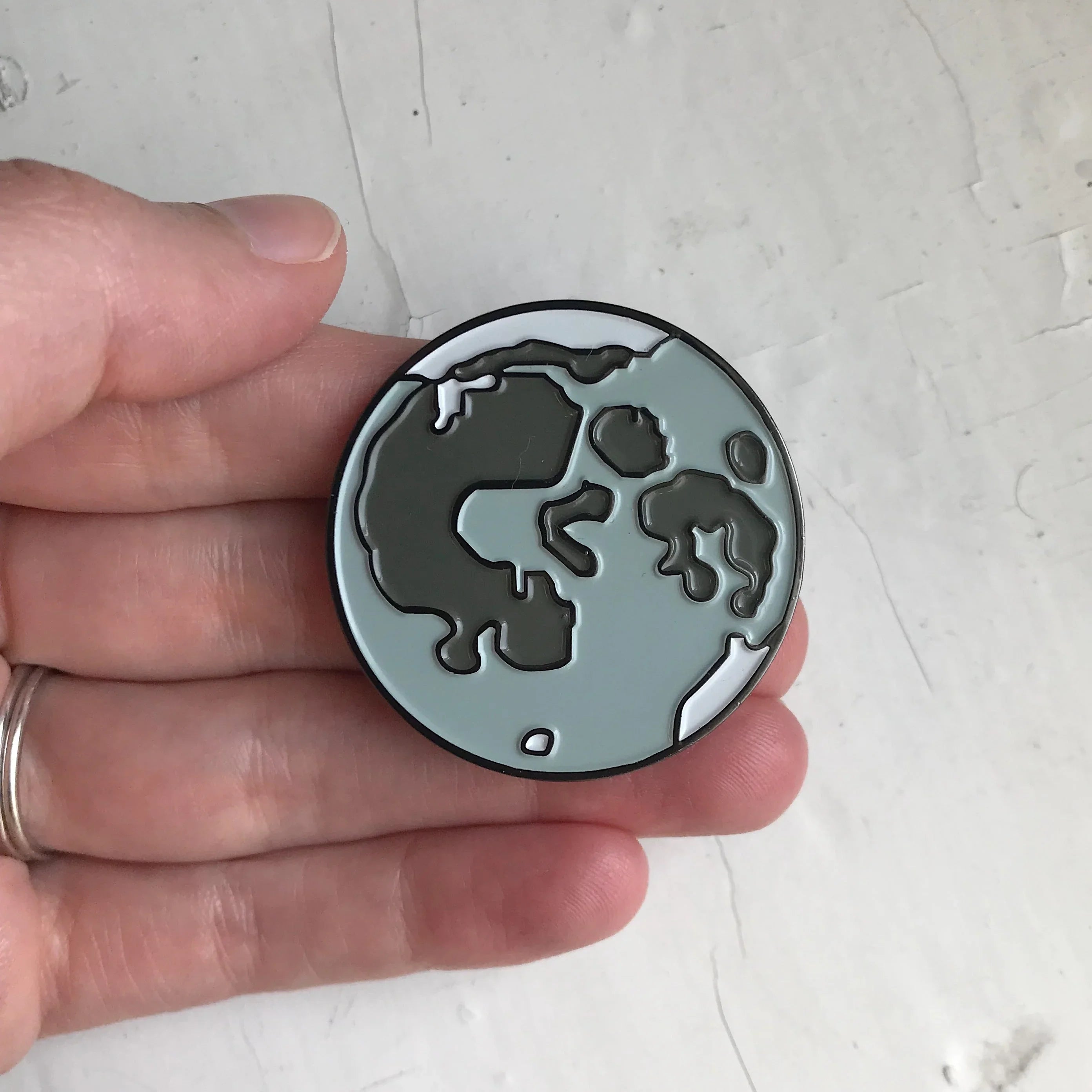 Hand holding a round enamel pin resembling the moon's cratered surface in gray tones