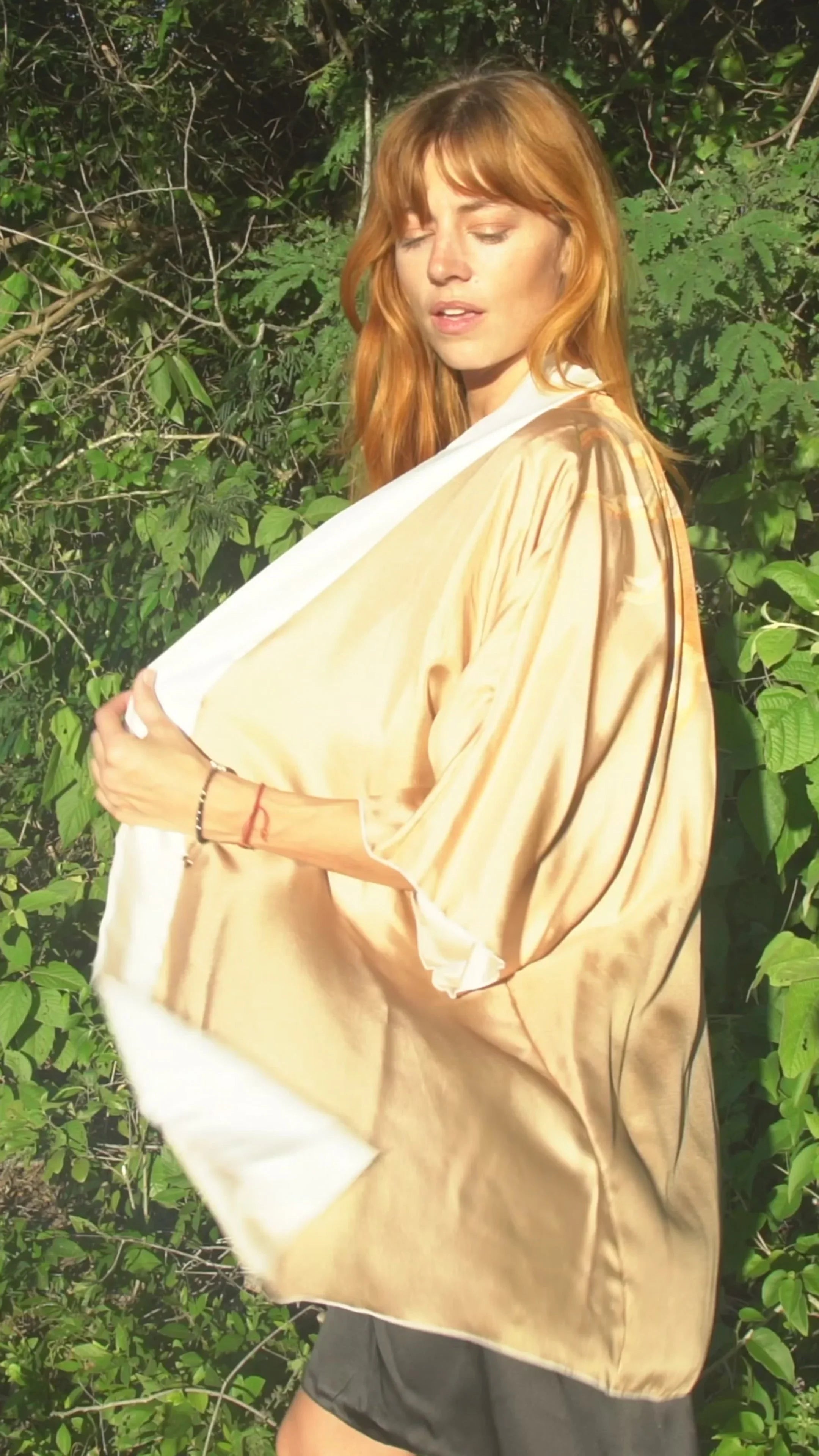 Woman with red hair wearing a beige satin shawl outdoors surrounded by green foliage