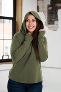 Smiling woman in an olive green hoodie from Qu!rk Boutique, standing in a modern loft.