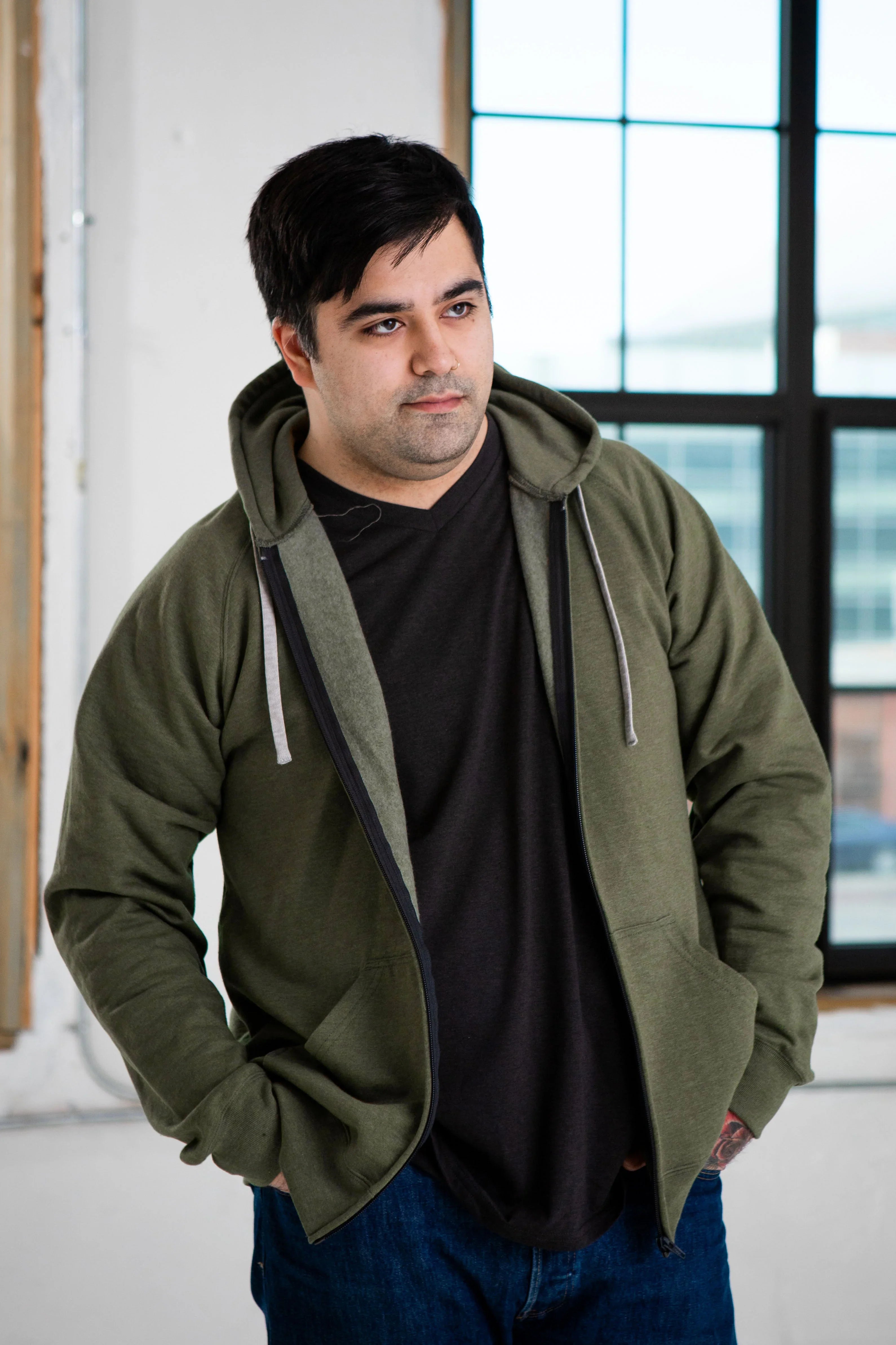 Man in olive hoodie and black t-shirt from Qu!rk Boutique, standing by window, casual style
