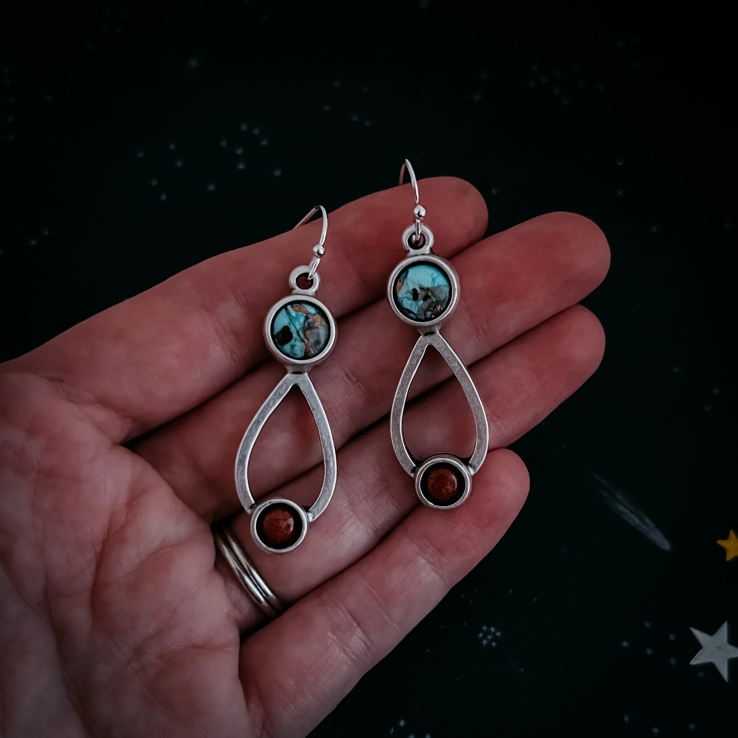 Hand holding silver earrings with turquoise and red coral stones on a dark star-patterned background