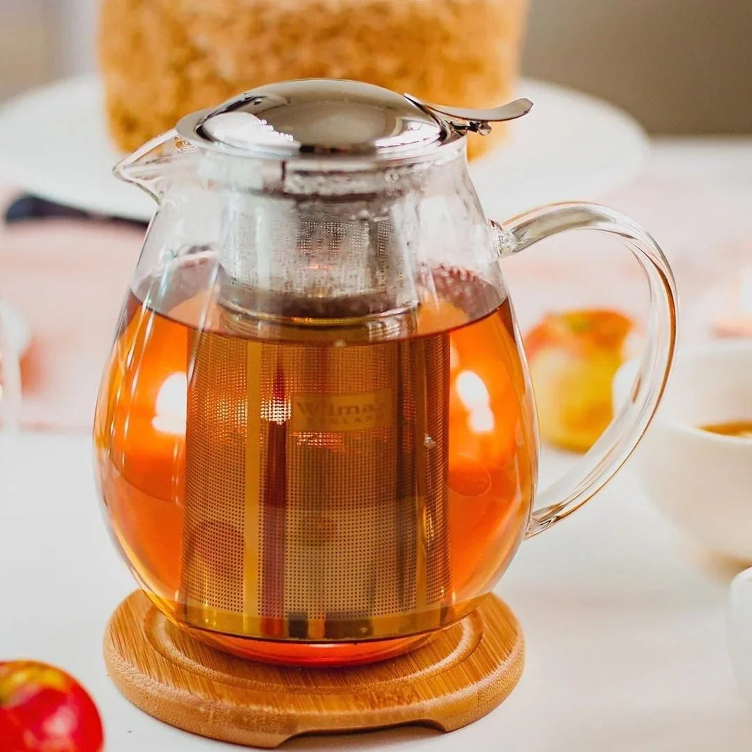 Glass teapot with metal infuser on bamboo coaster, filled with warm tea, cozy boutique setting