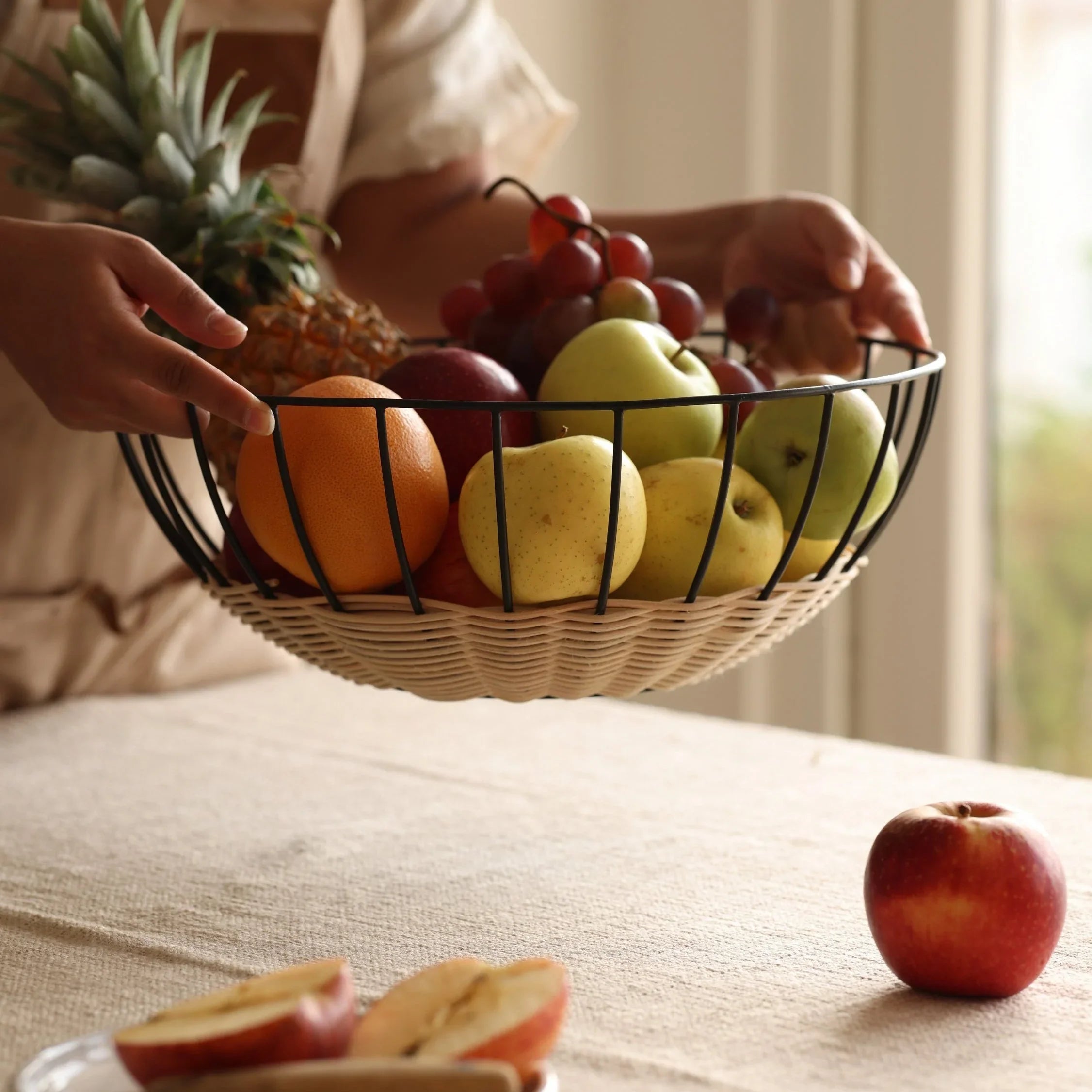 Hand holding a wire fruit basket with apples, grapes, orange, pineapple on a table at Qu!rk Boutique