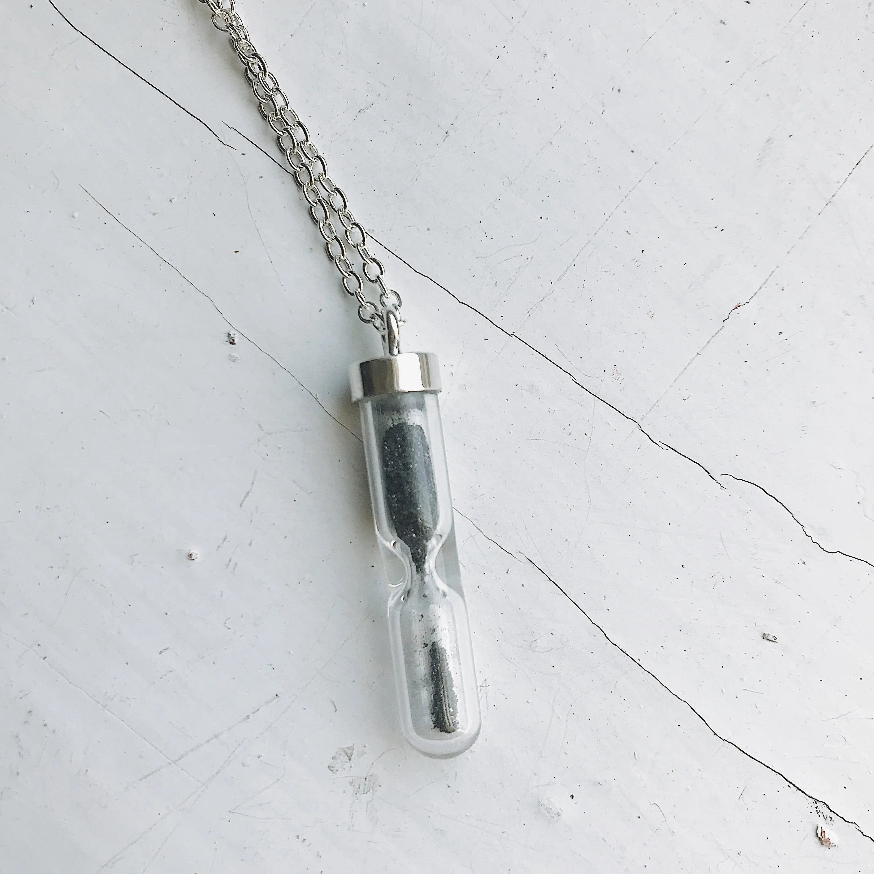 Silver chain necklace with a small transparent hourglass pendant filled with black sand on a cracked white surface