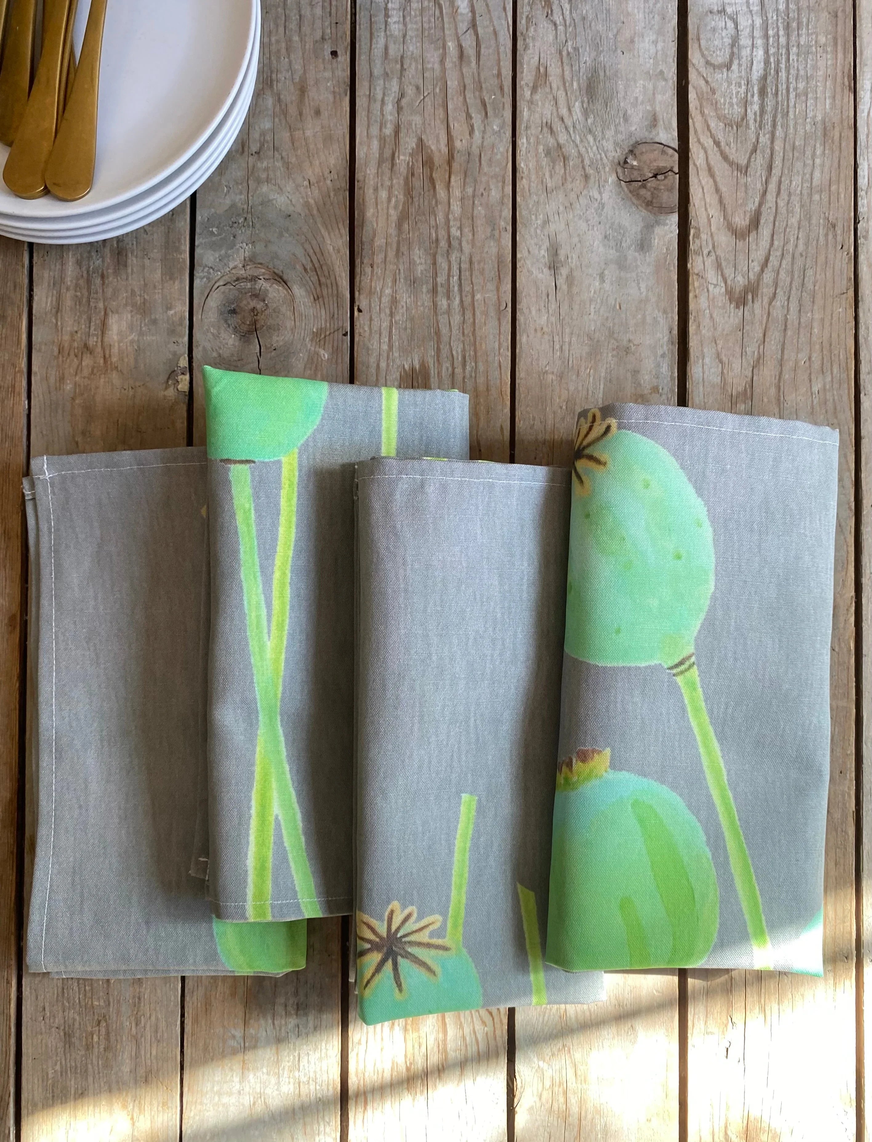Folded cotton napkins with green botanical print on rustic wooden table with stack of white plates and gold cutlery