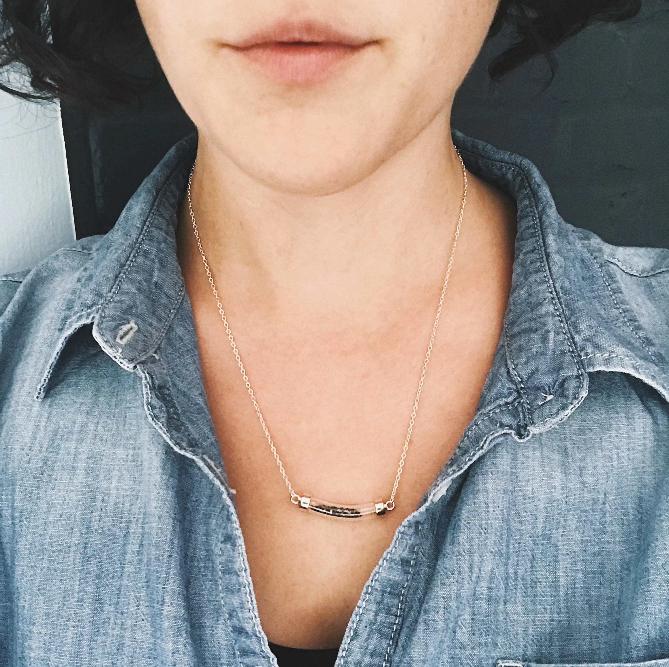 Close-up of a woman wearing a delicate gold bar necklace with a textured detail, dressed in a blue denim shirt
