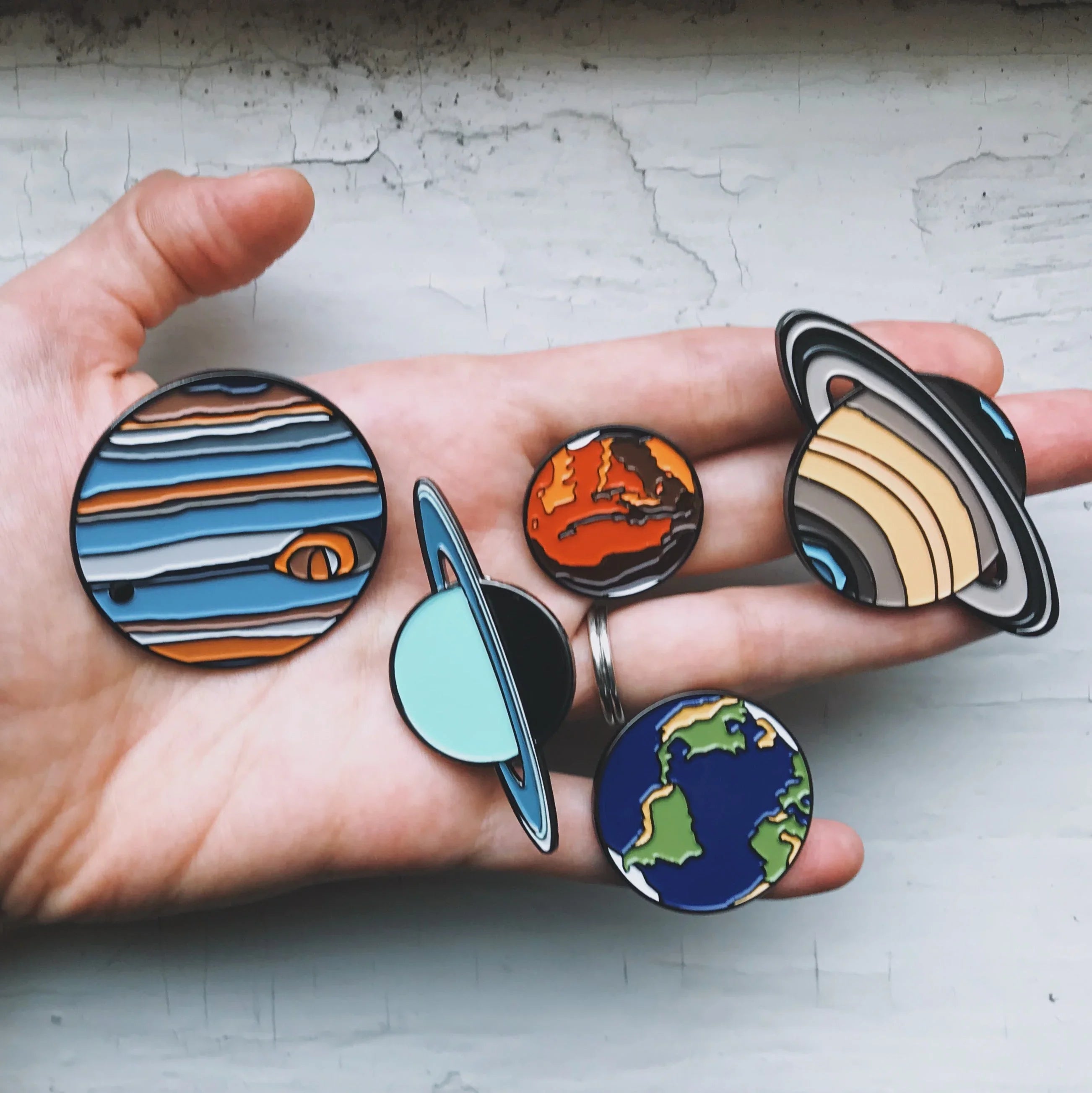Hand holding colorful enamel pins of planets Jupiter, Mars, Uranus, Earth, and Saturn on cracked white surface