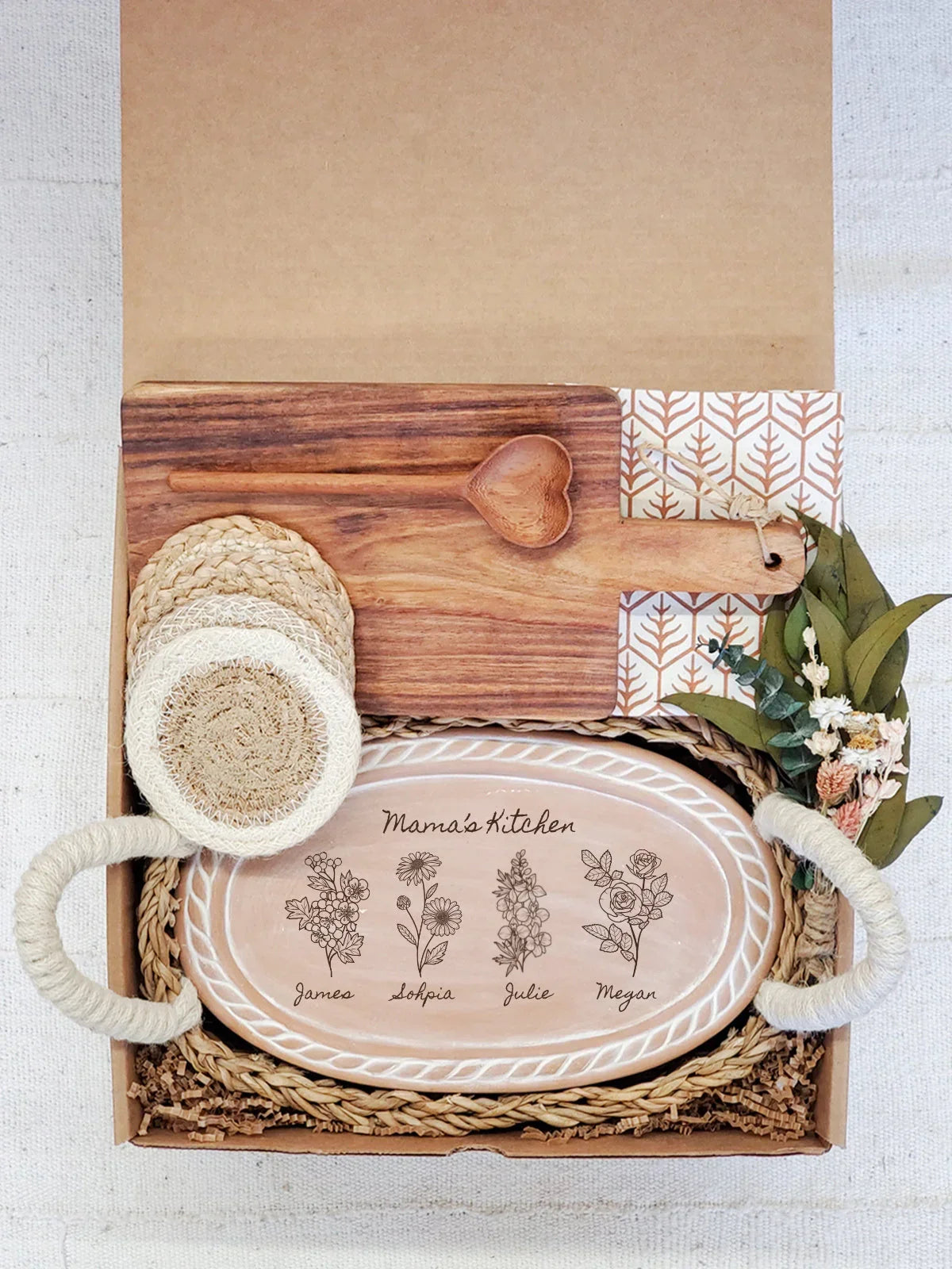 Gift box with wooden heart spoon, braided coasters, floral print plate labeled Mama's Kitchen, and dried flowers