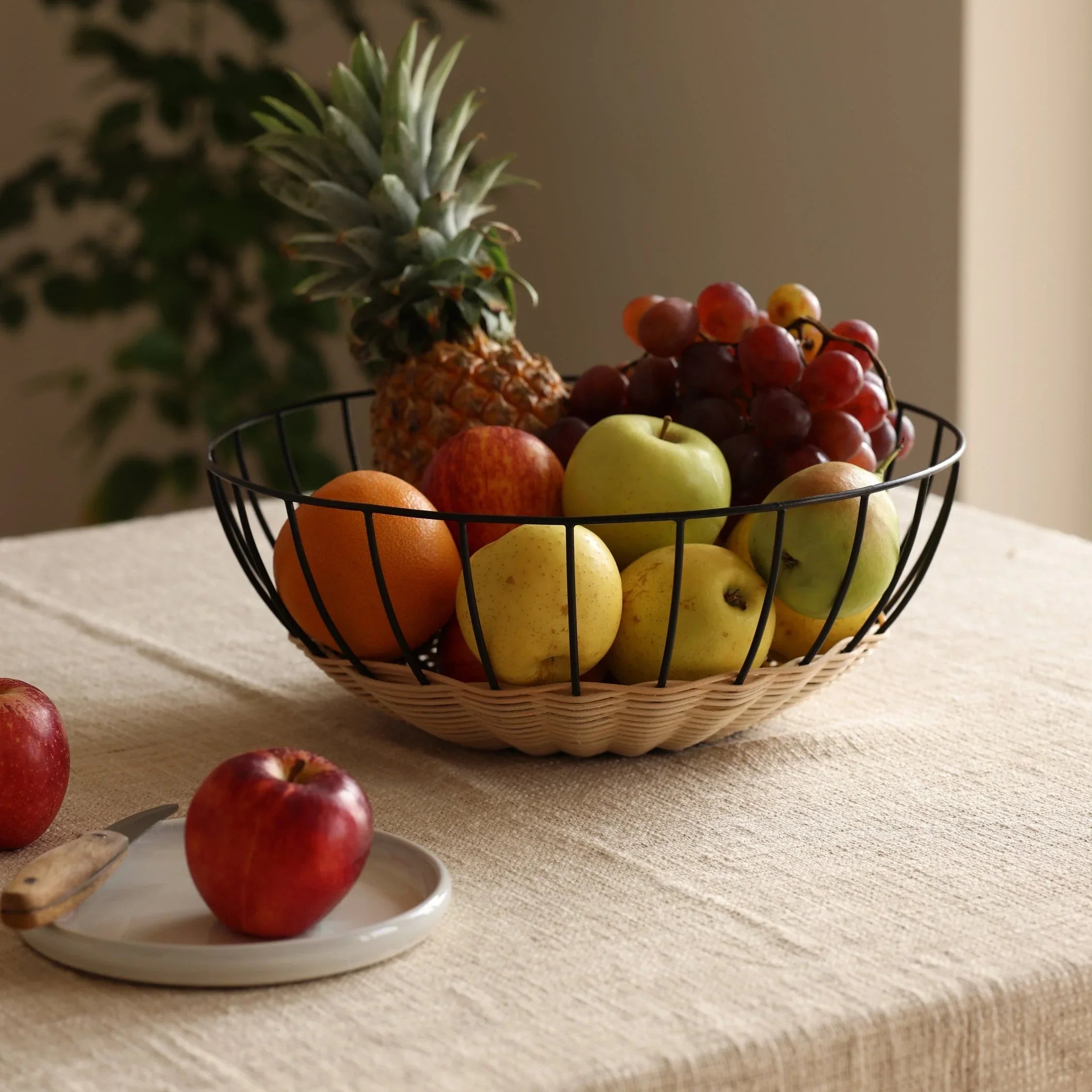 Wire fruit basket with pineapple, apples, oranges, grapes on neutral tablecloth, natural light