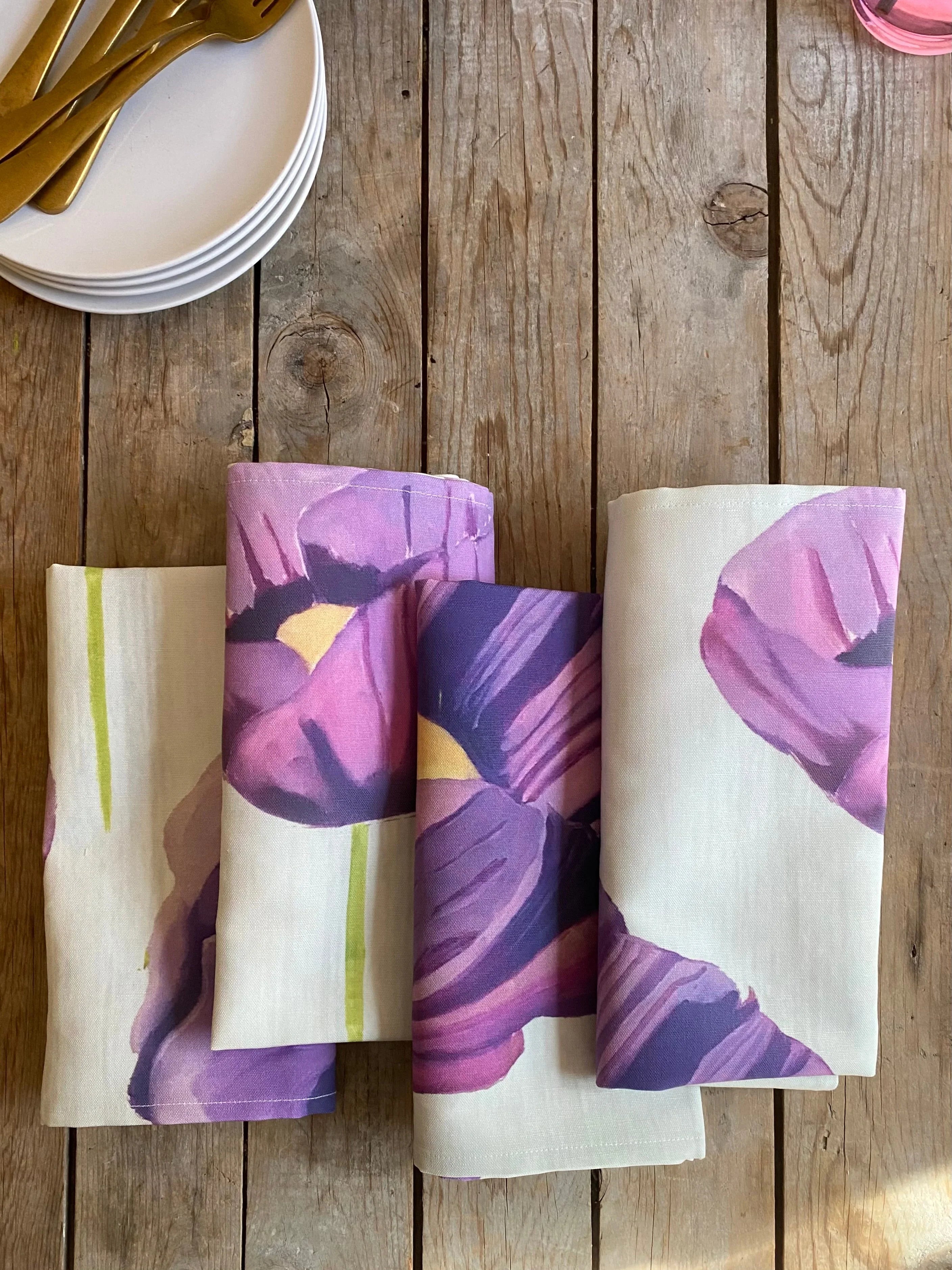 Folded purple floral cotton napkins on rustic wooden table with stack of white plates and gold cutlery