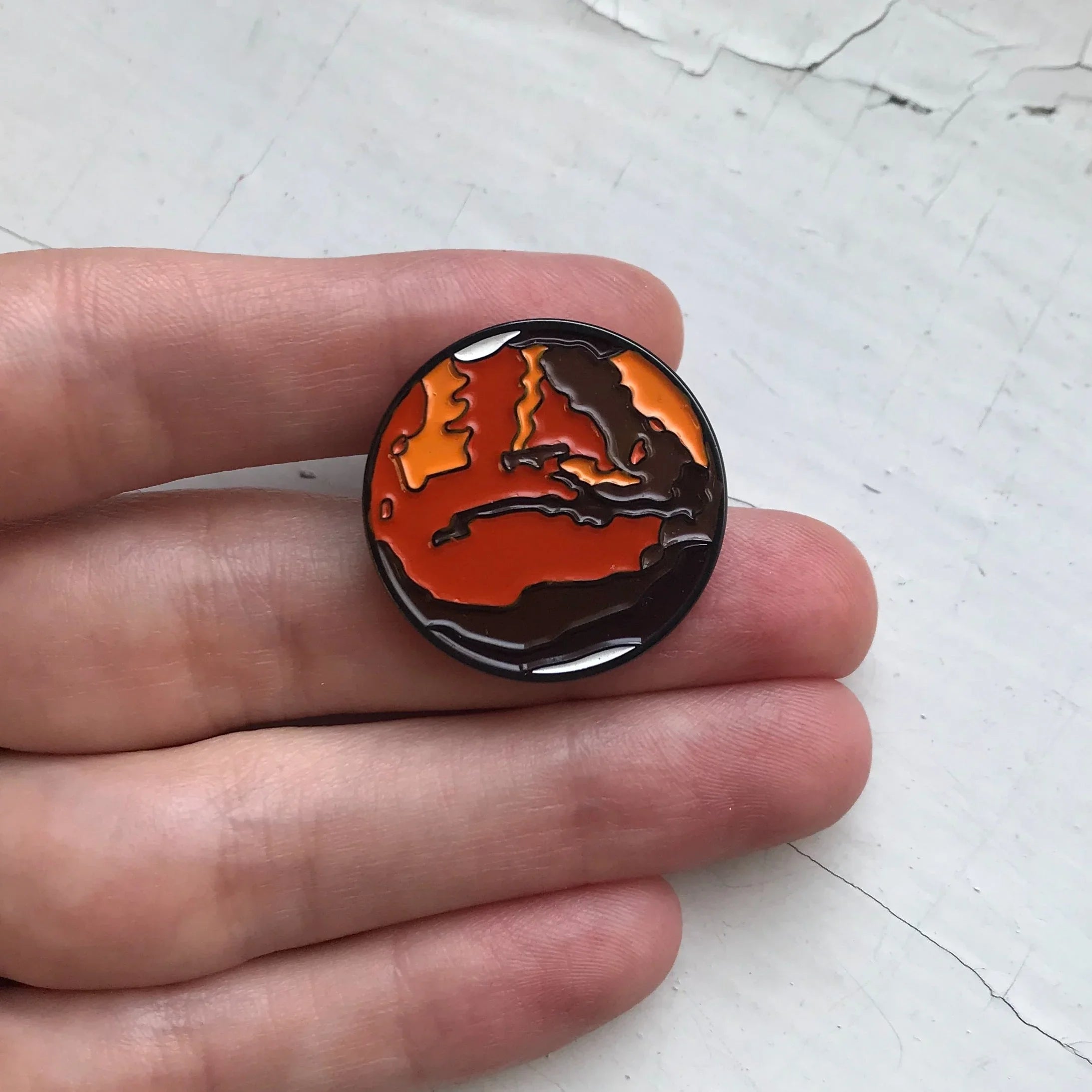 Hand holding an enamel pin featuring a stylized orange and brown map design on a white textured surface