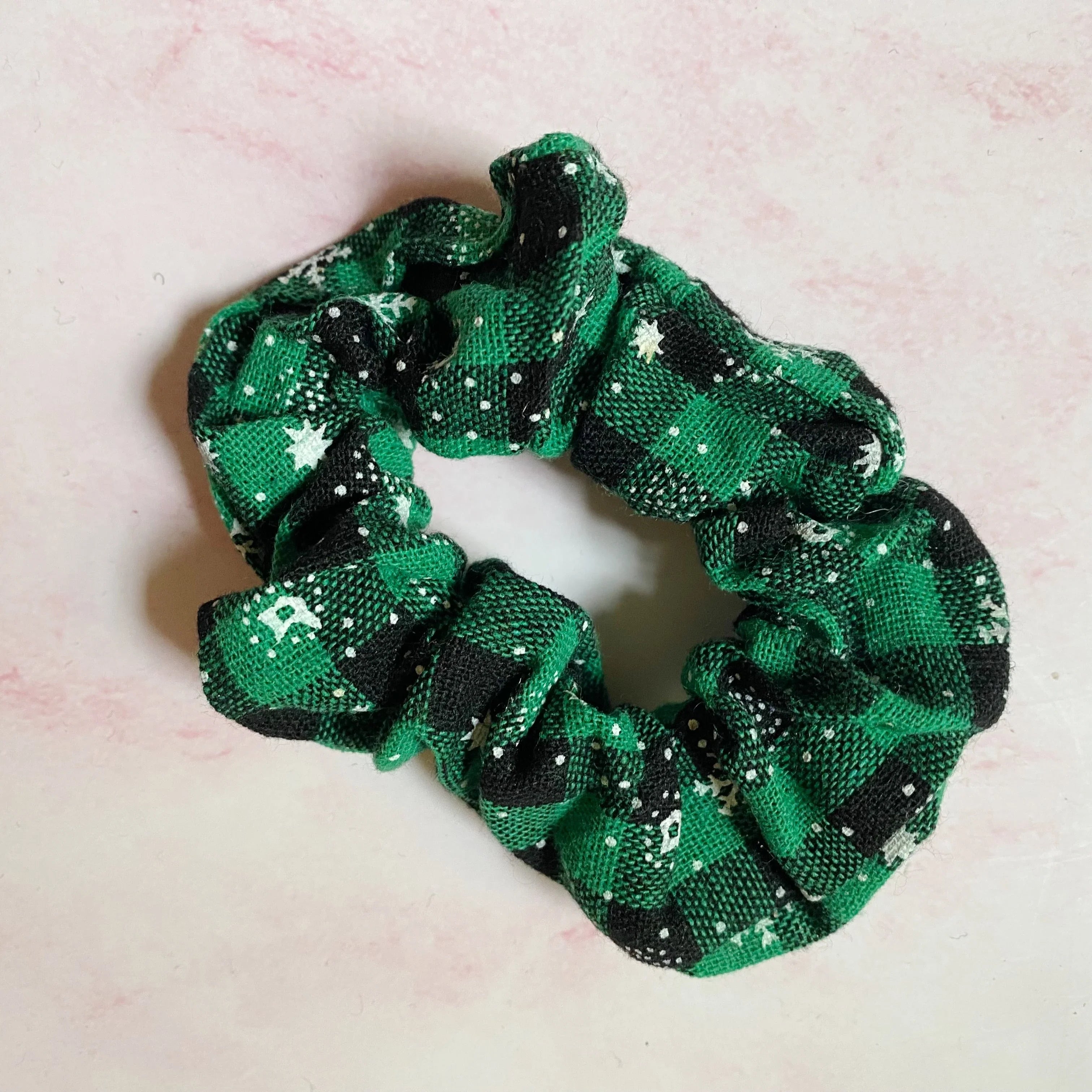 Green and black plaid fabric scrunchie with white star and snowflake patterns on pink background