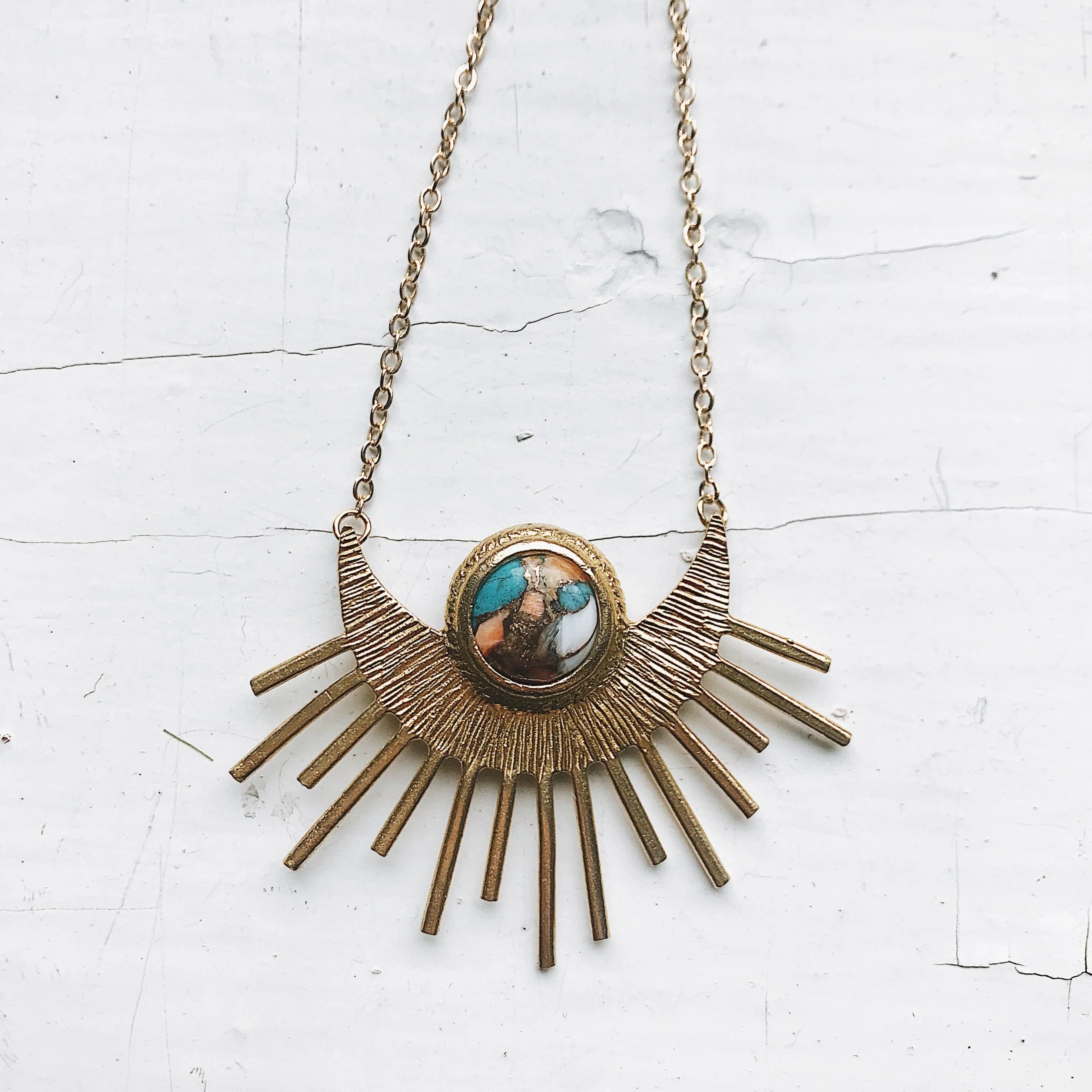 Gold metal sunburst pendant necklace with turquoise and brown stone centerpiece on white cracked surface