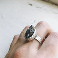 Hand wearing a hammered silver ring featuring a rough, dark meteorite stone on a white background