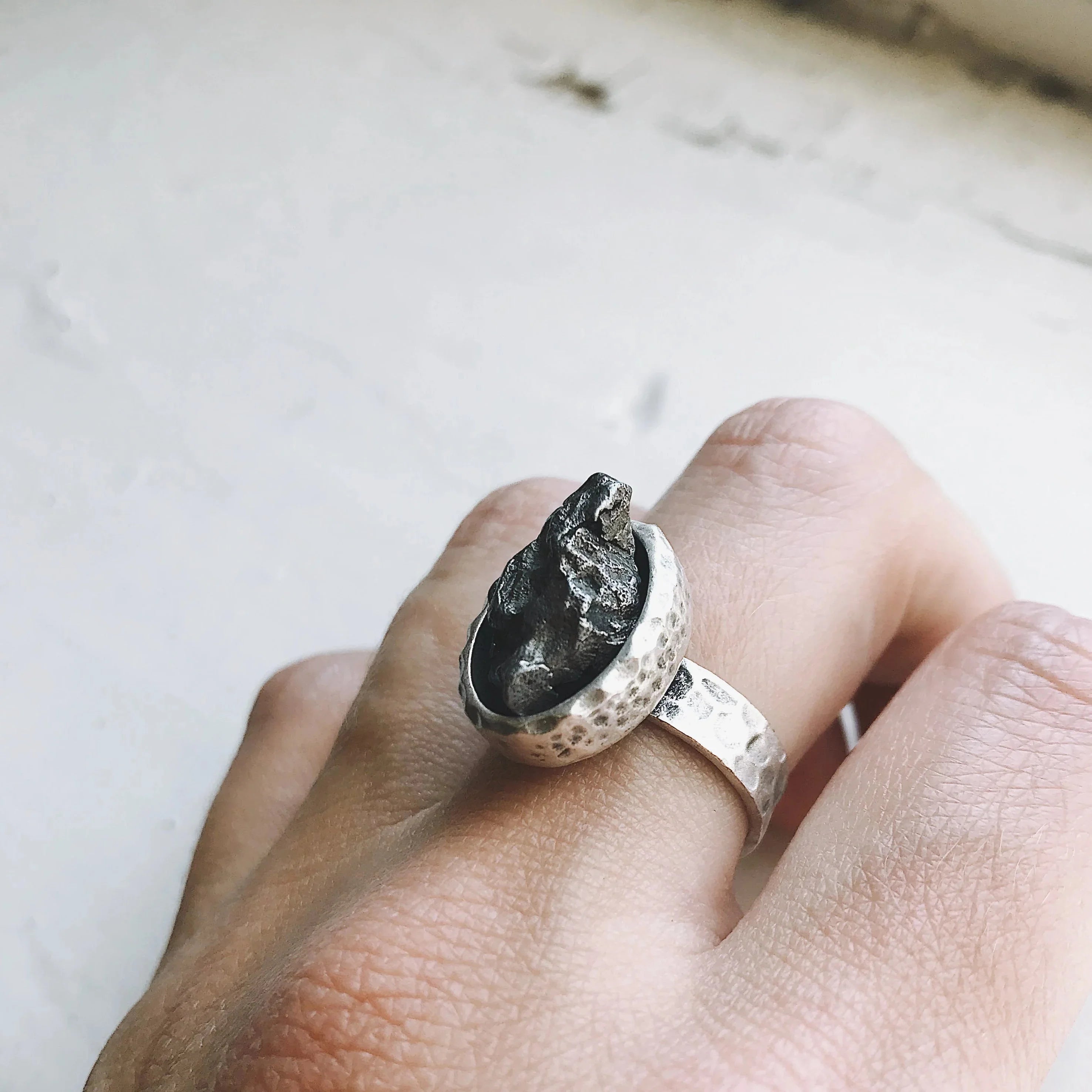 Hand wearing a hammered silver ring featuring a rough, dark meteorite stone on a white background