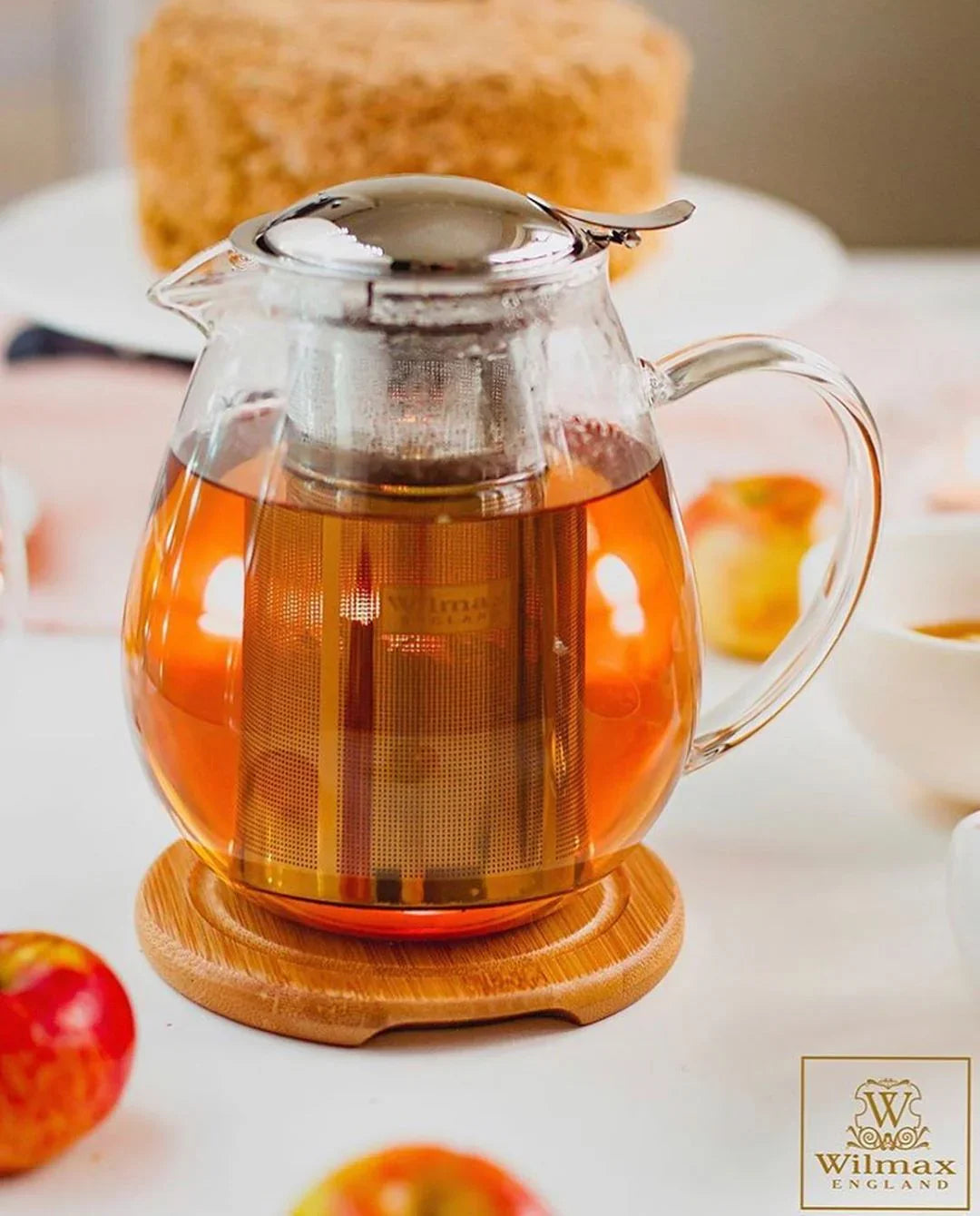 Clear glass teapot with metal infuser, filled with tea, on bamboo coaster, apples nearby on table