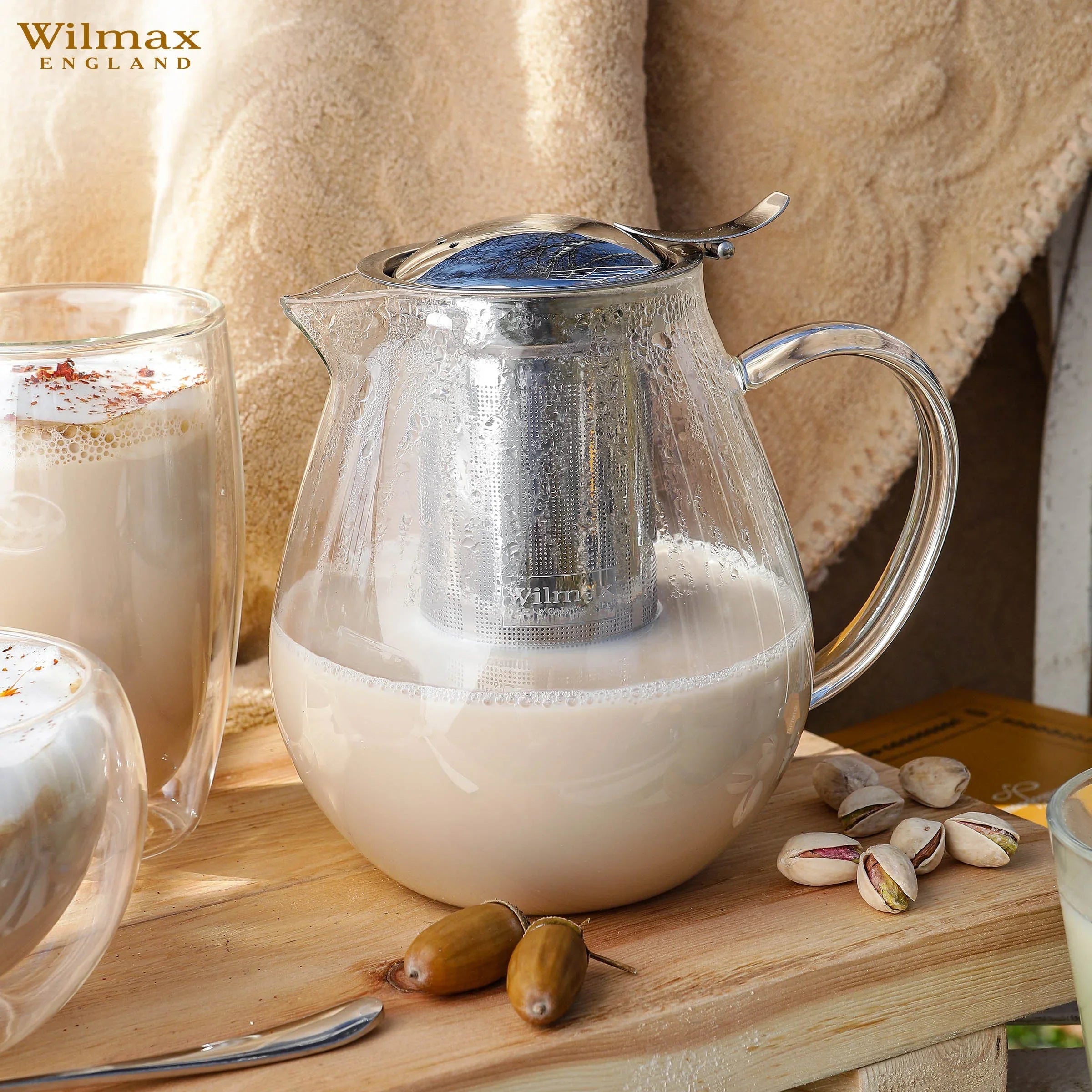 Glass teapot with stainless steel infuser brewing tea latte, surrounded by pistachios and mugs on wood table