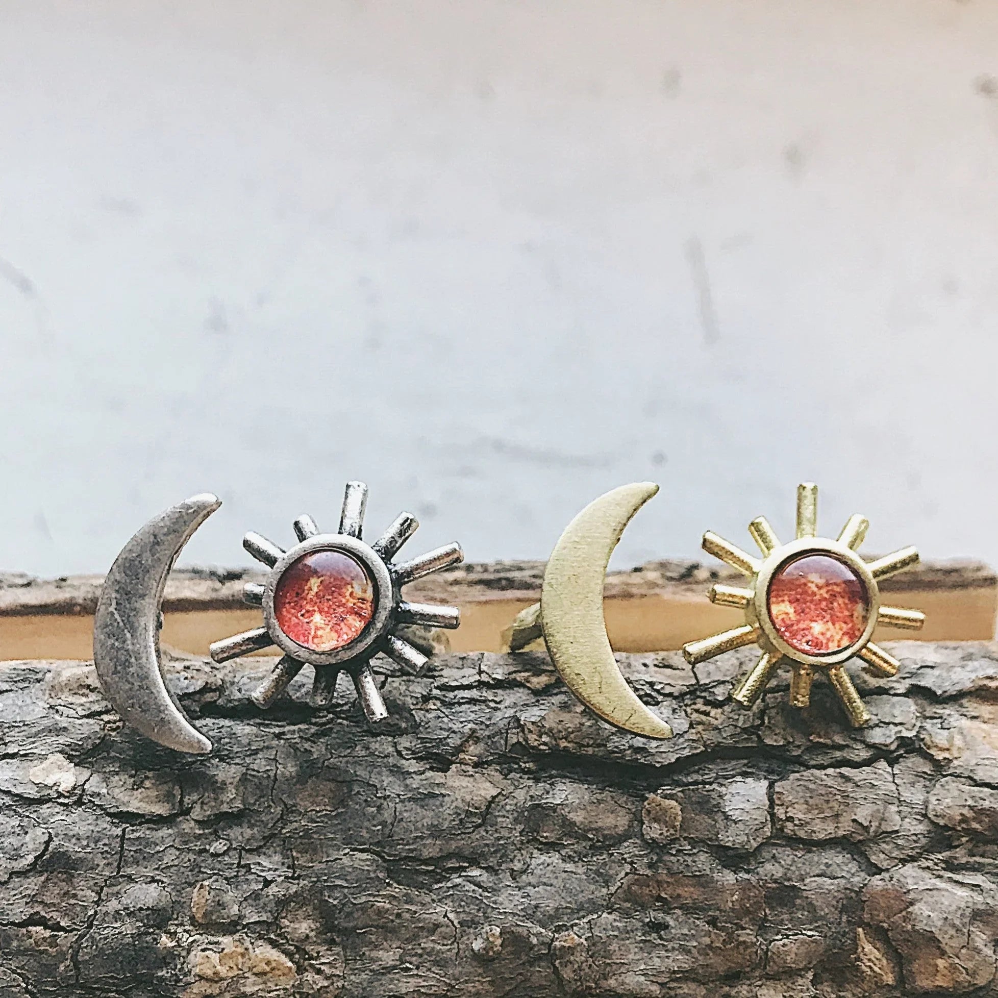 Two pairs of sun and crescent moon earrings on textured wooden surface, silver and gold tones