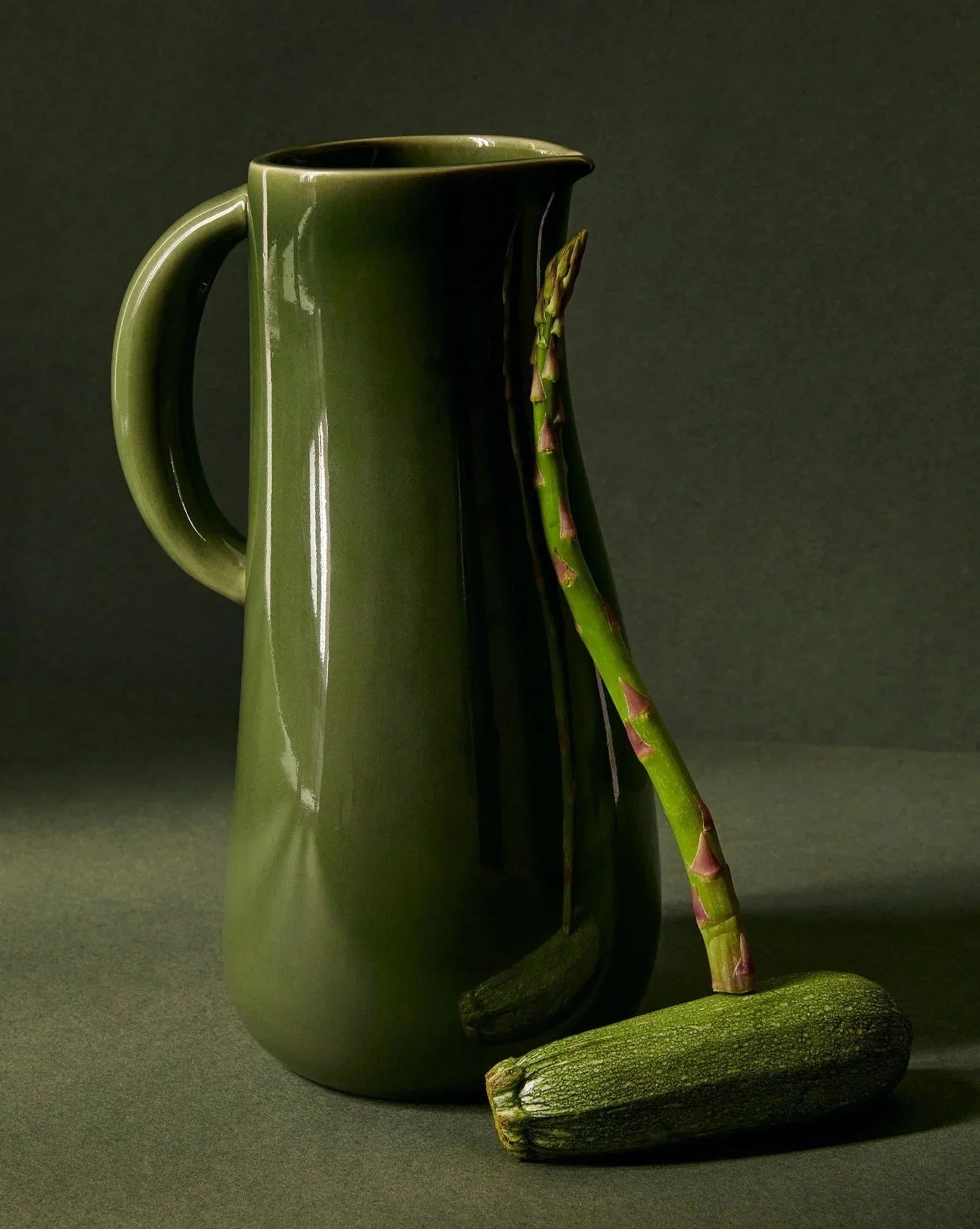 Green ceramic pitcher with an asparagus spear and zucchini, styled on a dark backdrop at Qu!rk Boutique