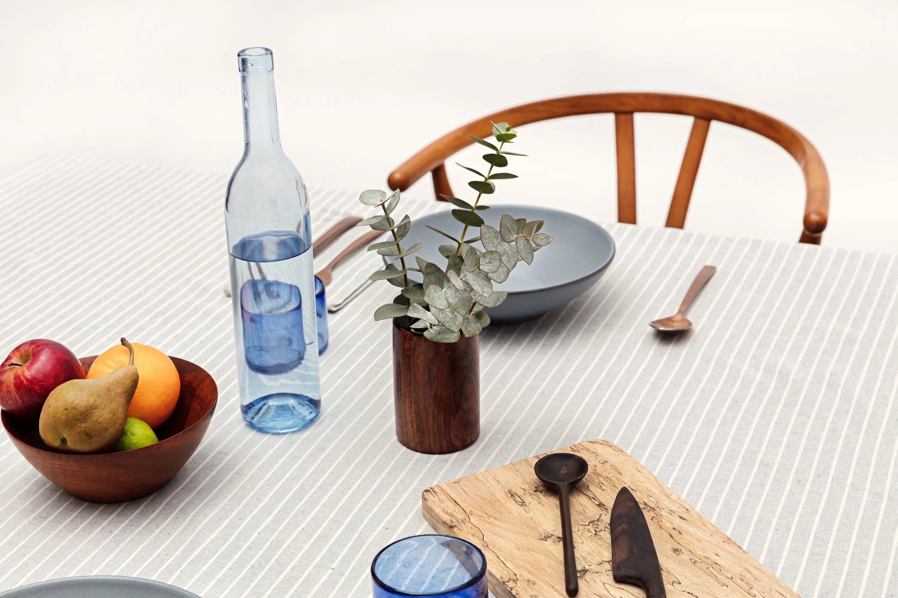 Minimalist table setting with striped tablecloth, wooden chair, fruit bowl, blue glassware, and eucalyptus in vase.