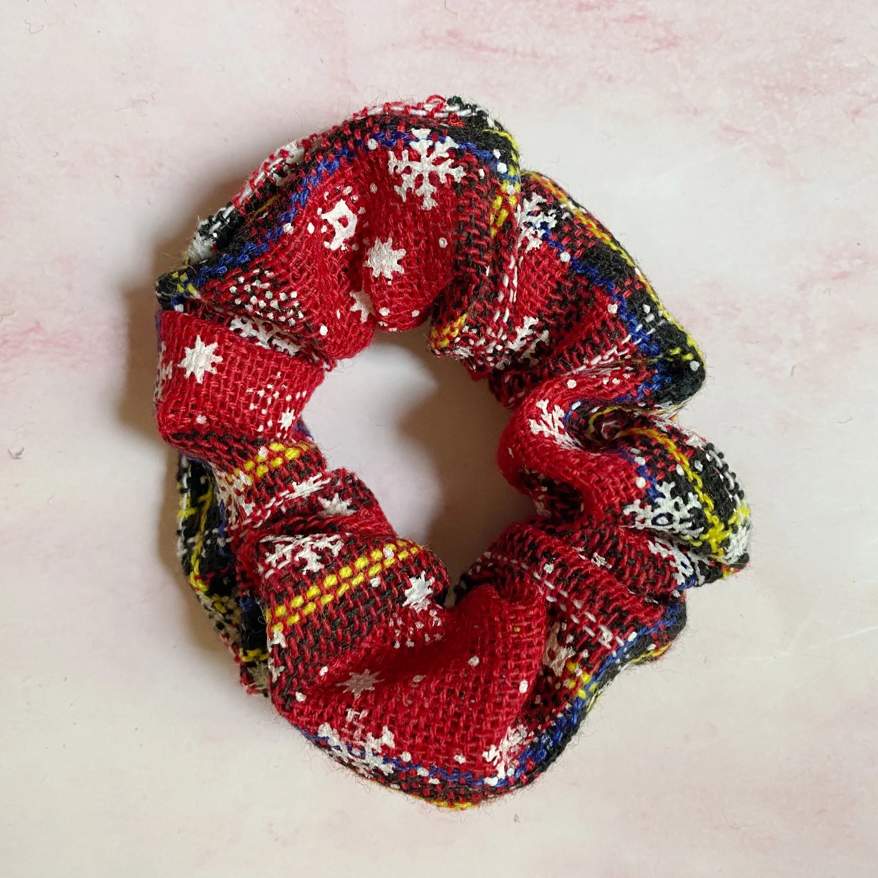 Red woven fabric scrunchie with white snowflake and plaid patterns on light pink surface