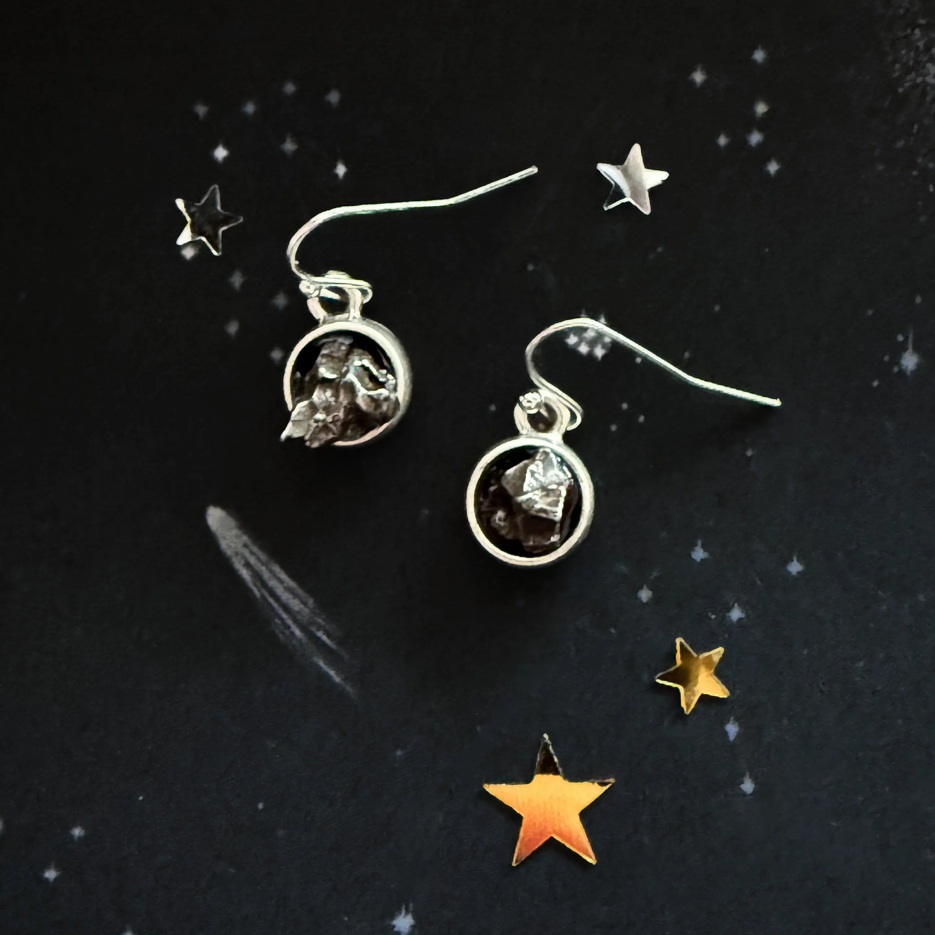 Silver dangle earrings with raw black stones on a black starry background with gold and silver stars