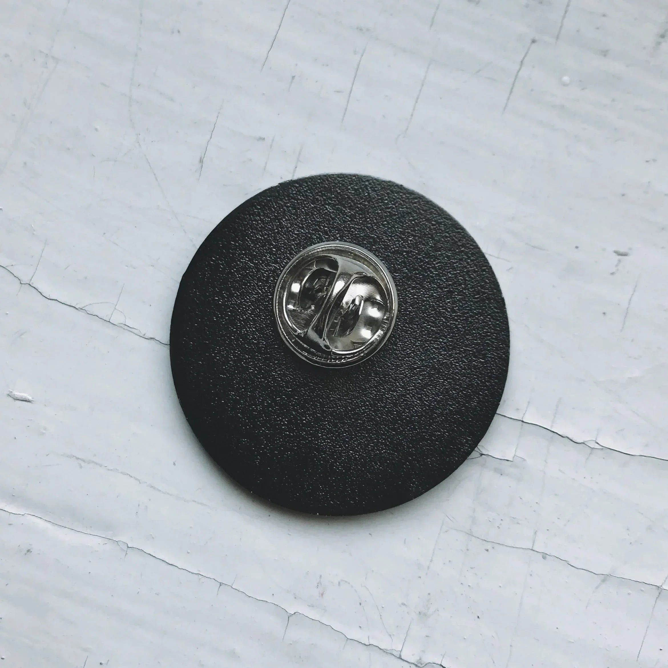 Close-up of black enamel pin back with silver butterfly clutch on cracked white surface