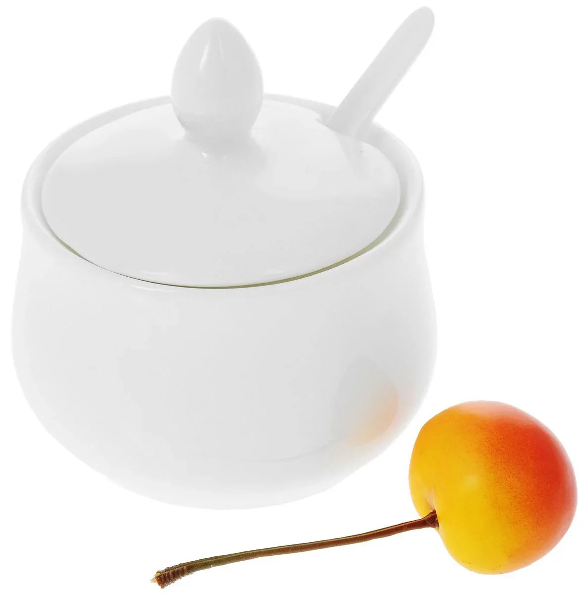 White ceramic sugar bowl with lid and spoon, next to a yellow-red cherry, on white background.