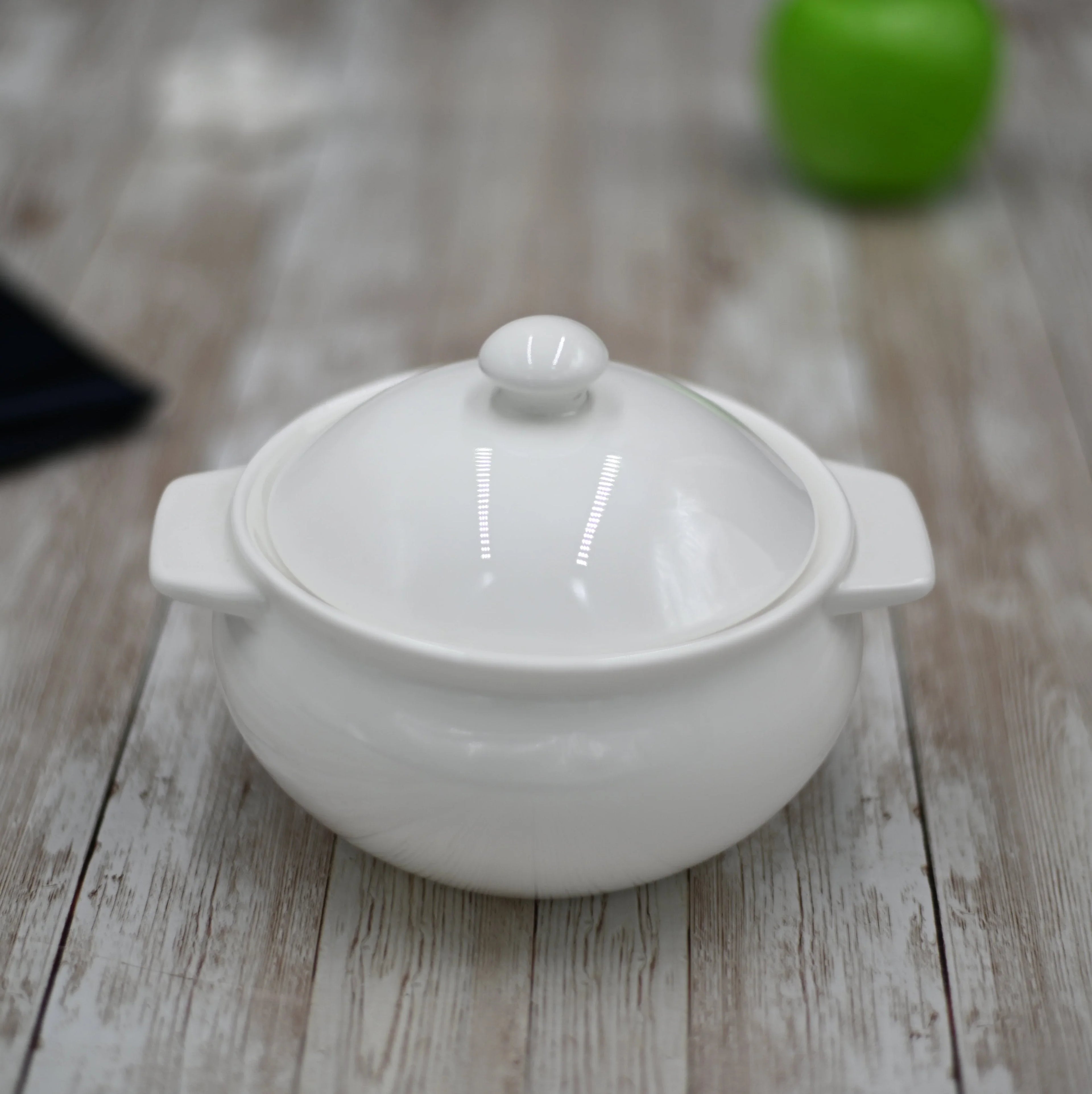 White ceramic pot with lid on rustic wooden table, green apple blurred in background
