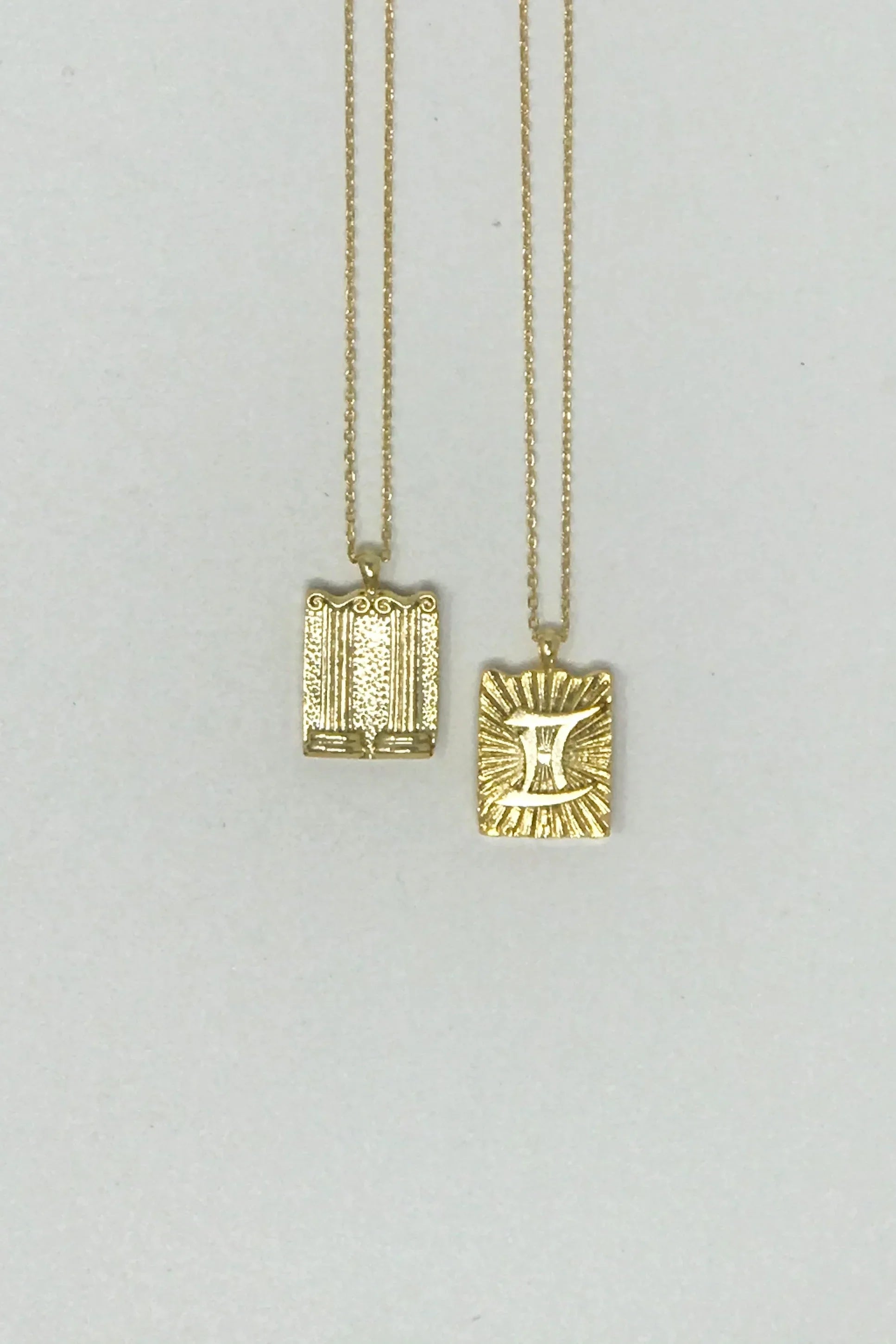 Two gold rectangular pendant necklaces on chains, one with Gemini zodiac symbol and rays, the other with decorative columns