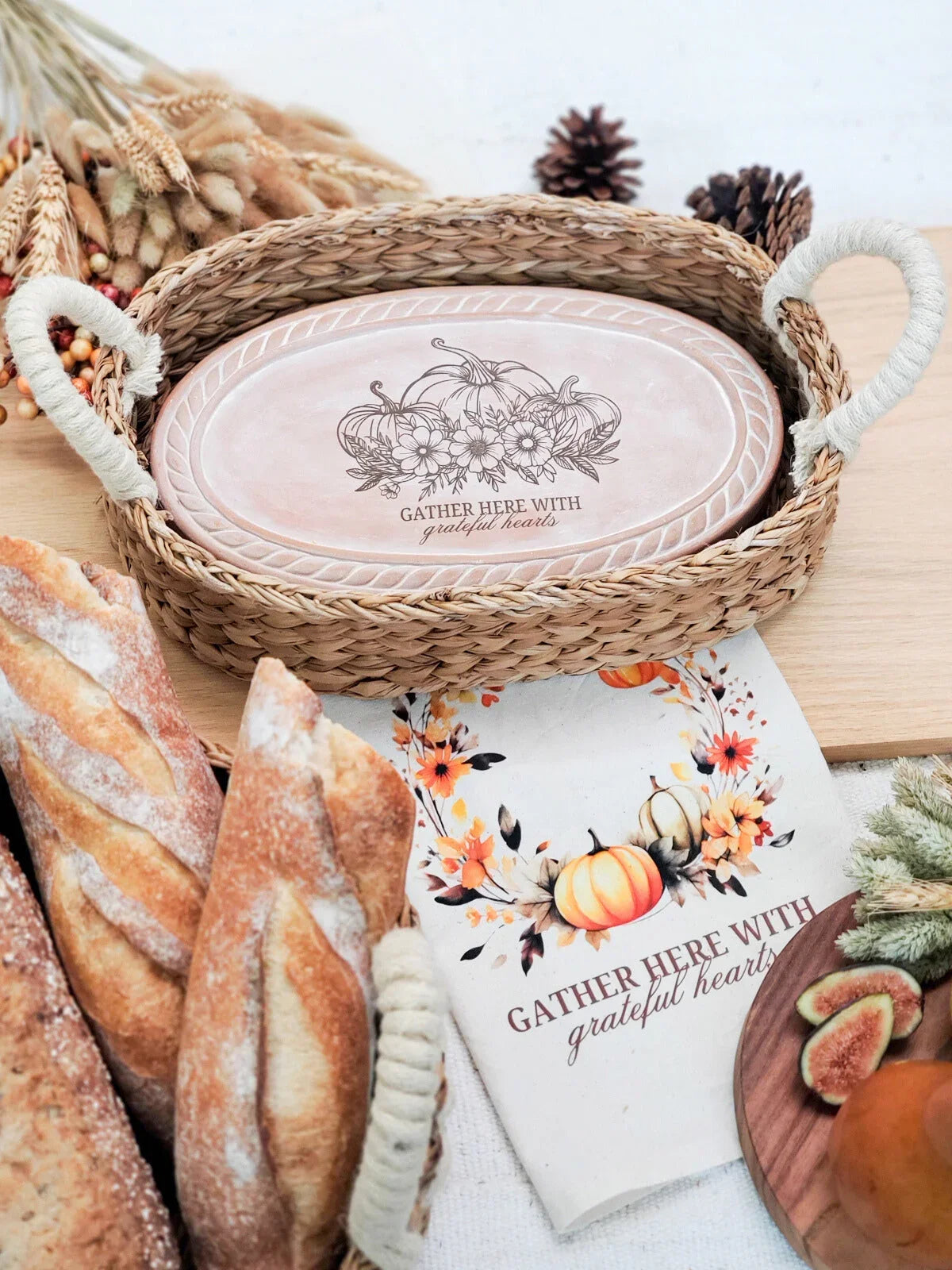 woven basket with rustic tray saying 'Gather here with grateful hearts' surrounded by bread, dried florals, pine cones, and a fall-themed cloth