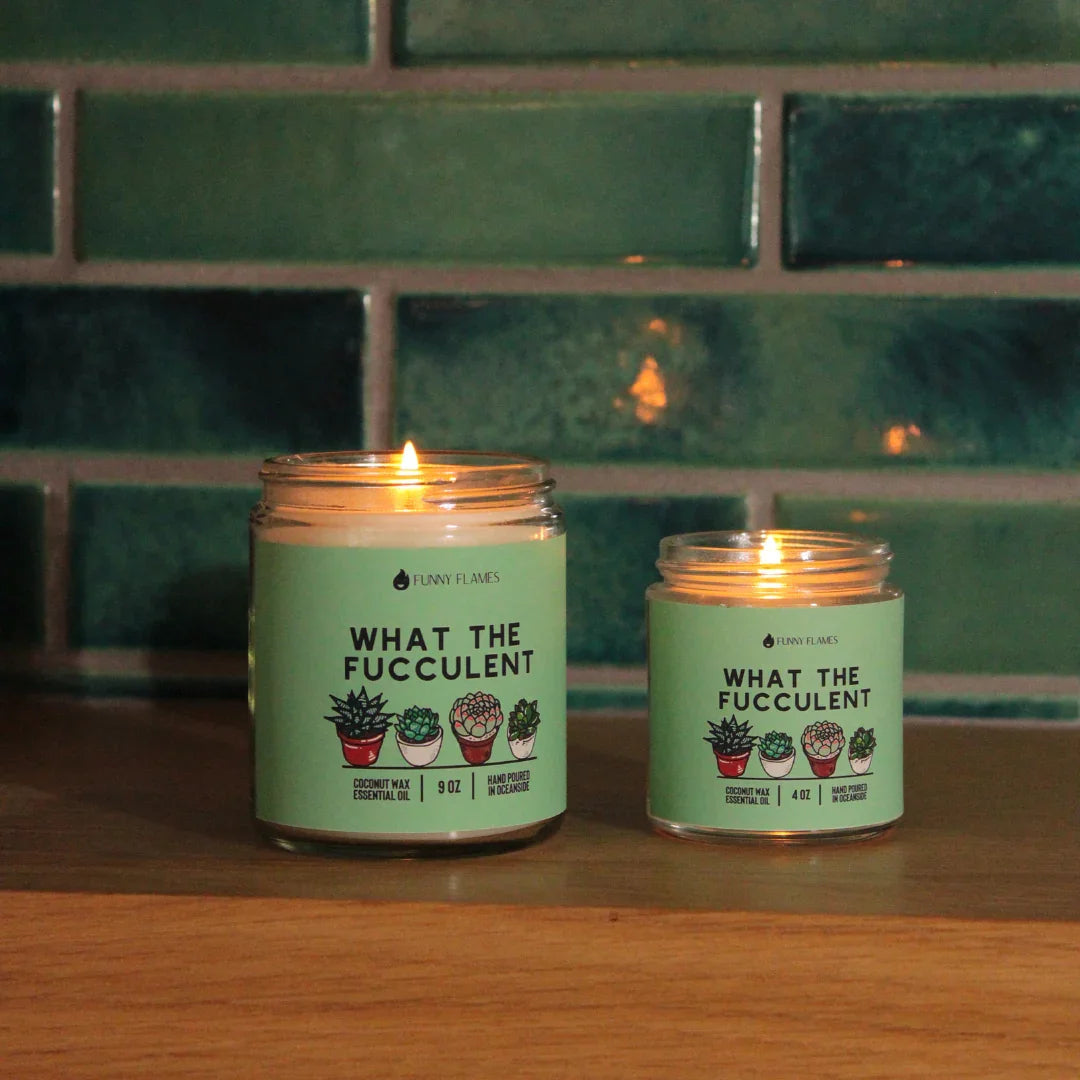 Two lit scented candles labeled What The Fucculent with succulent plant illustrations on wooden surface
