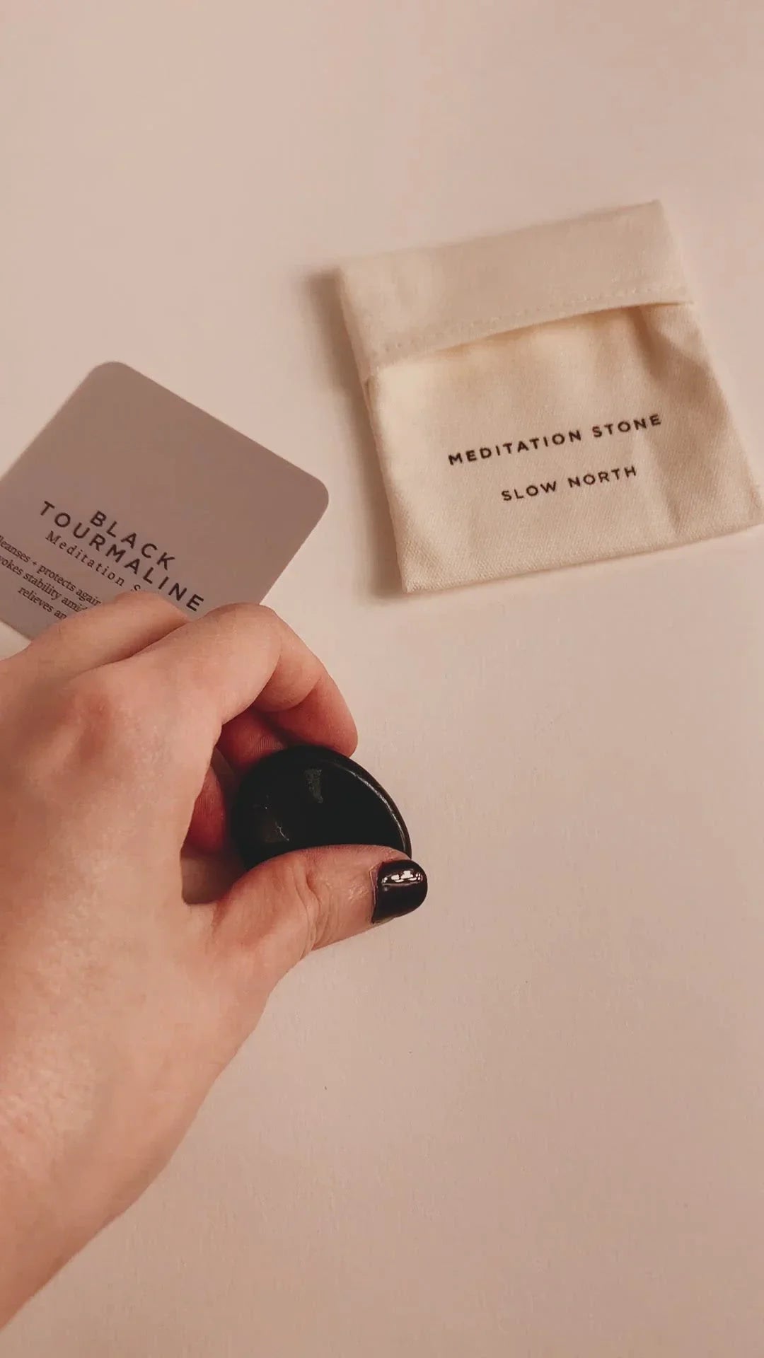 Hand holding smooth black tourmaline meditation stone with Slow North pouch and info card on beige surface