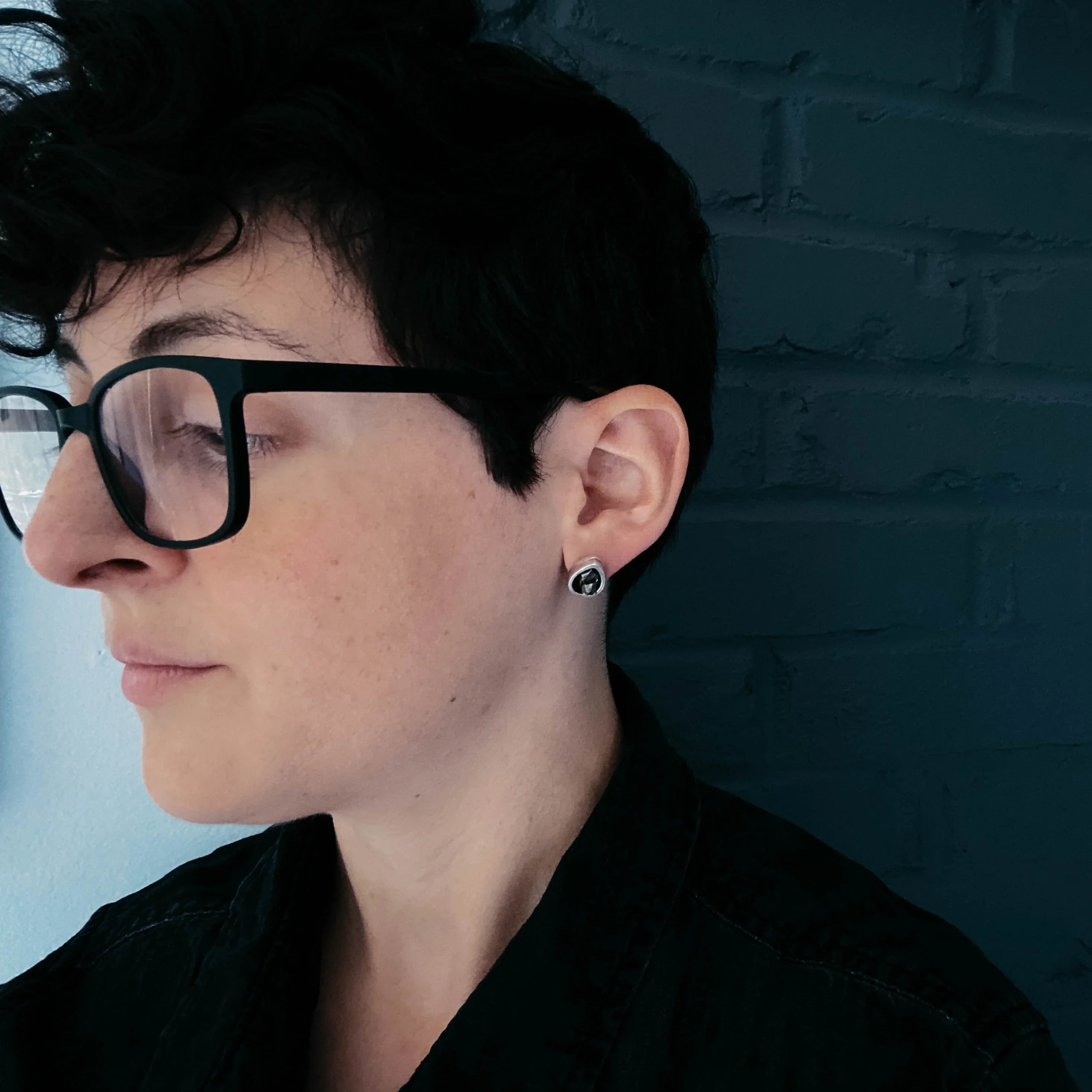 Person wearing black glasses and an artist-designed earring against a dark brick wall