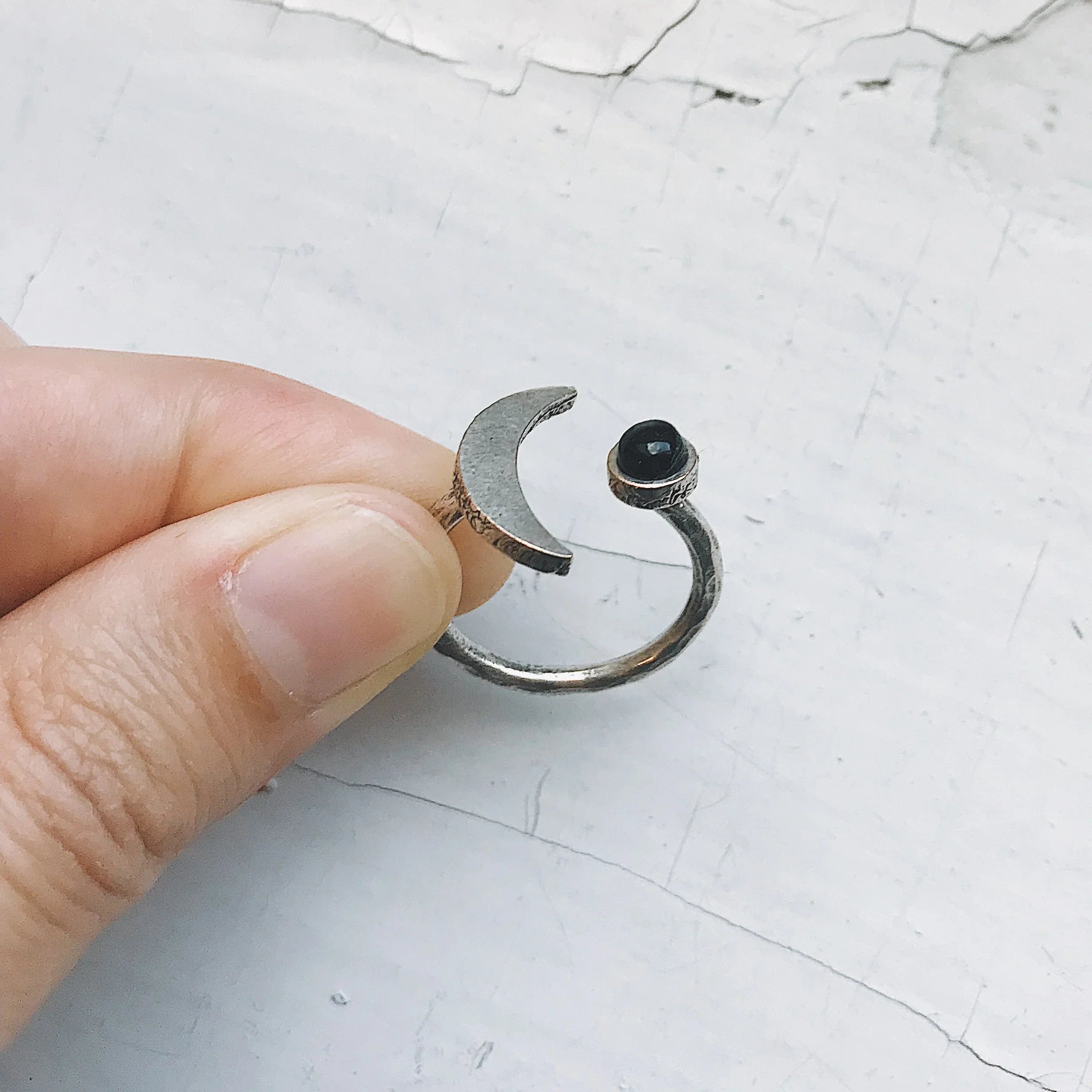 Hand holding silver adjustable ring with crescent moon and black stone on cracked white surface