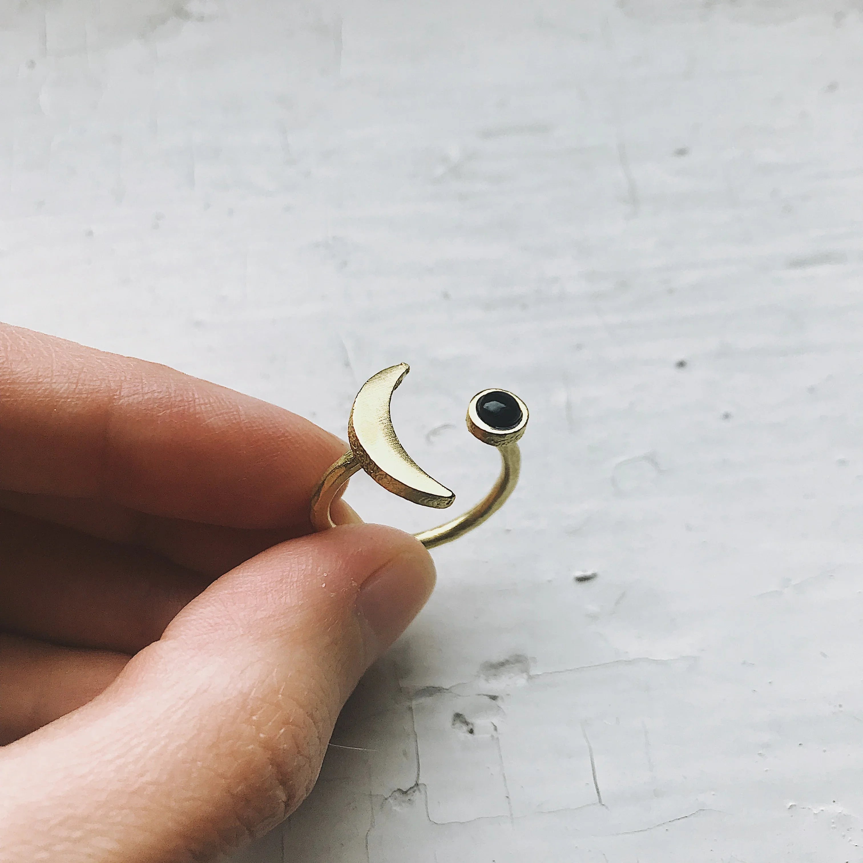 Hand holding adjustable gold ring with crescent moon and black stone designs on textured white background