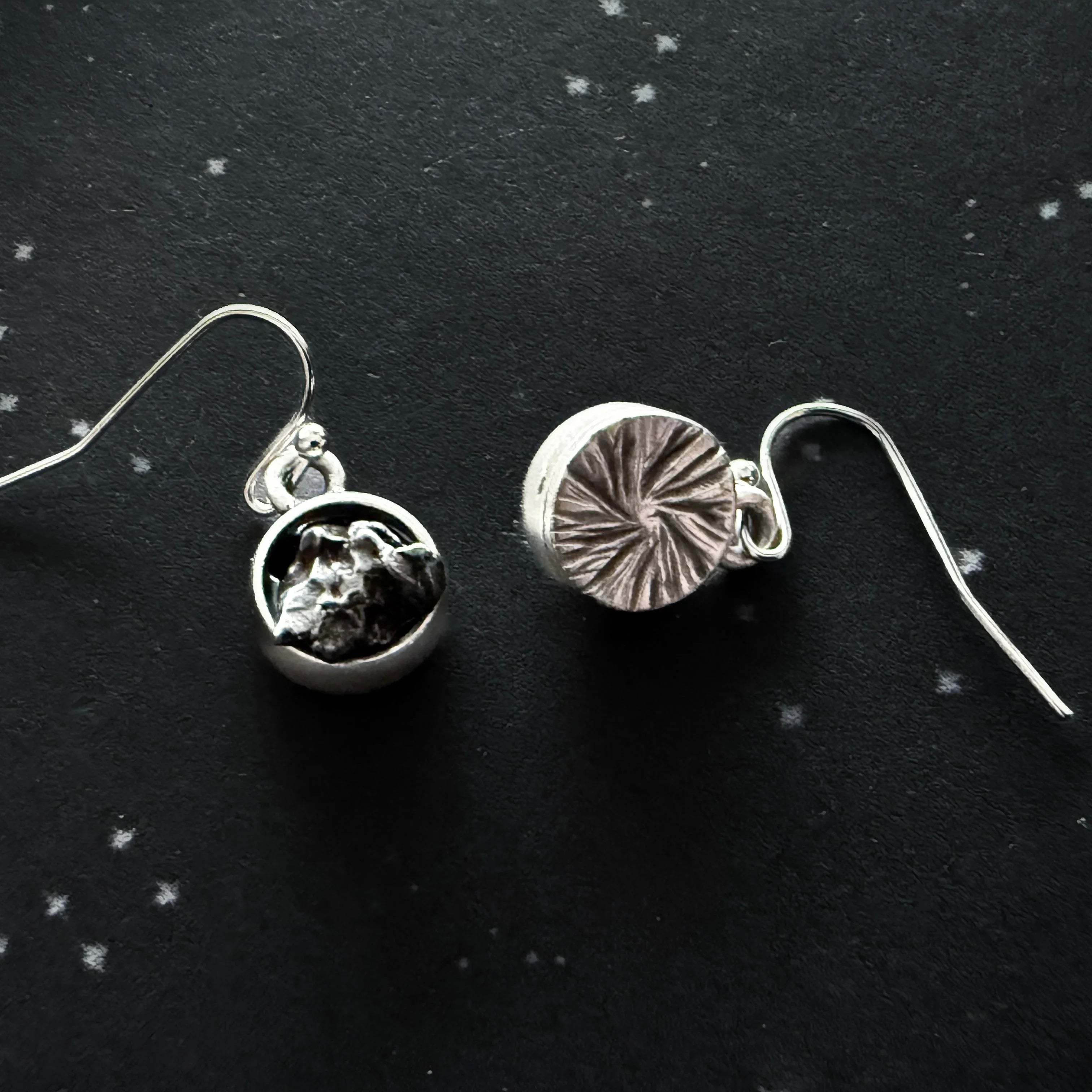Pair of silver hook earrings with textured circular pendants on black speckled background