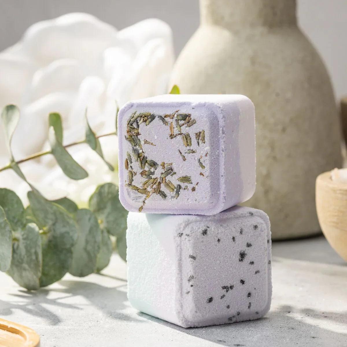 Two lavender and pale blue bath bombs stacked with dried herbs, eucalyptus, and a vase in the background