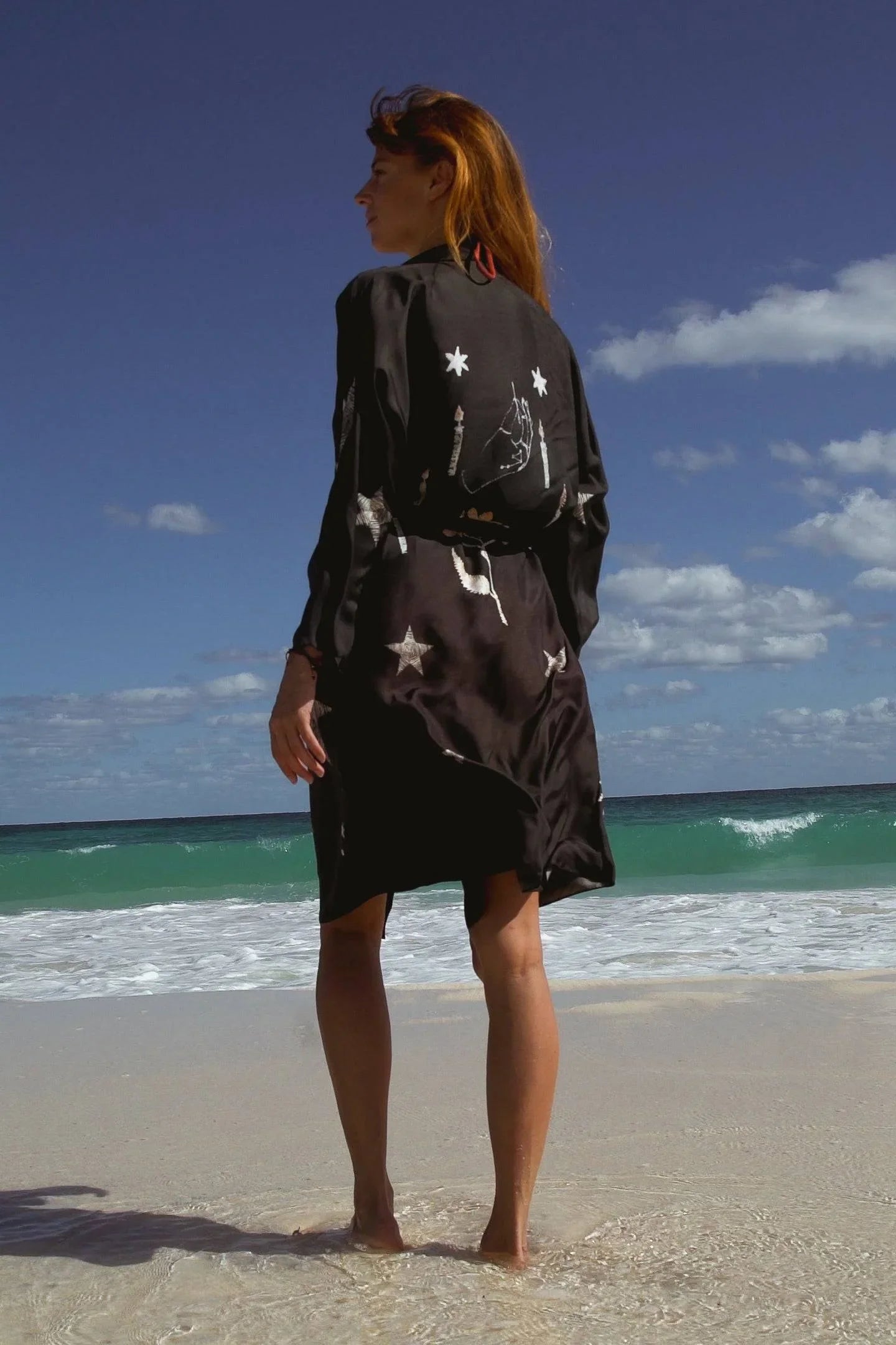 Woman with red hair wearing black dress with star patterns standing barefoot on beach by ocean