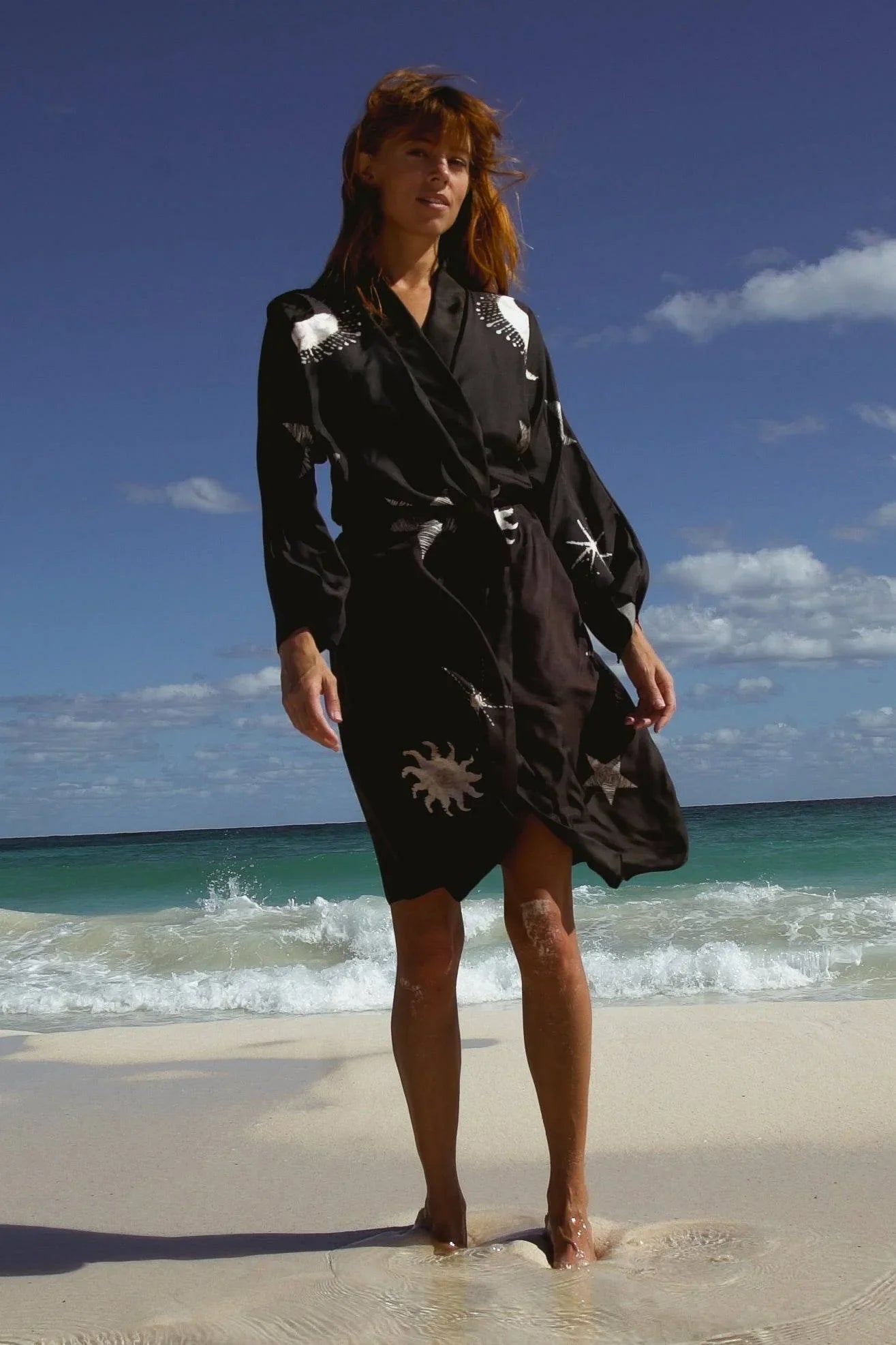 Woman standing barefoot on sandy beach wearing black robe with sun and star patterns under blue sky
