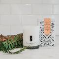 Amber Noir candle by tlc candle co. with decorative box, fern, and bark on white tile background