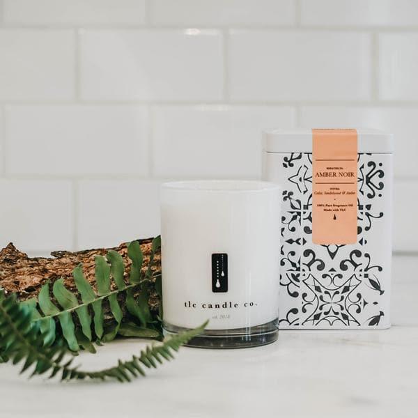 Amber Noir candle by tlc candle co. with decorative box, fern, and bark on white tile background
