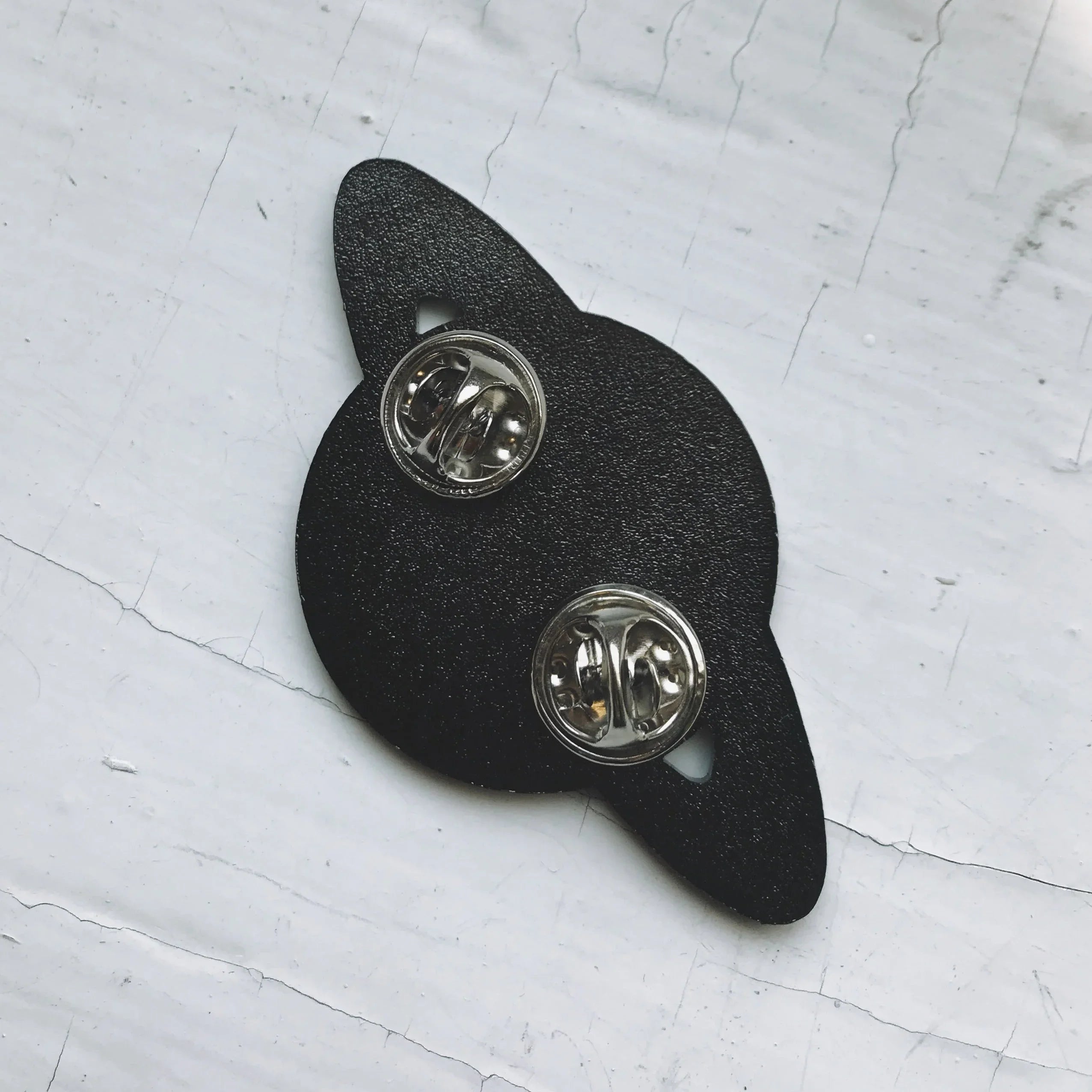 Back of a black planet-shaped enamel pin with two metal clasps on a white surface