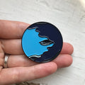 Hand holding an artist-designed enamel pin with a blue and navy cosmic face, Qu!rk Boutique