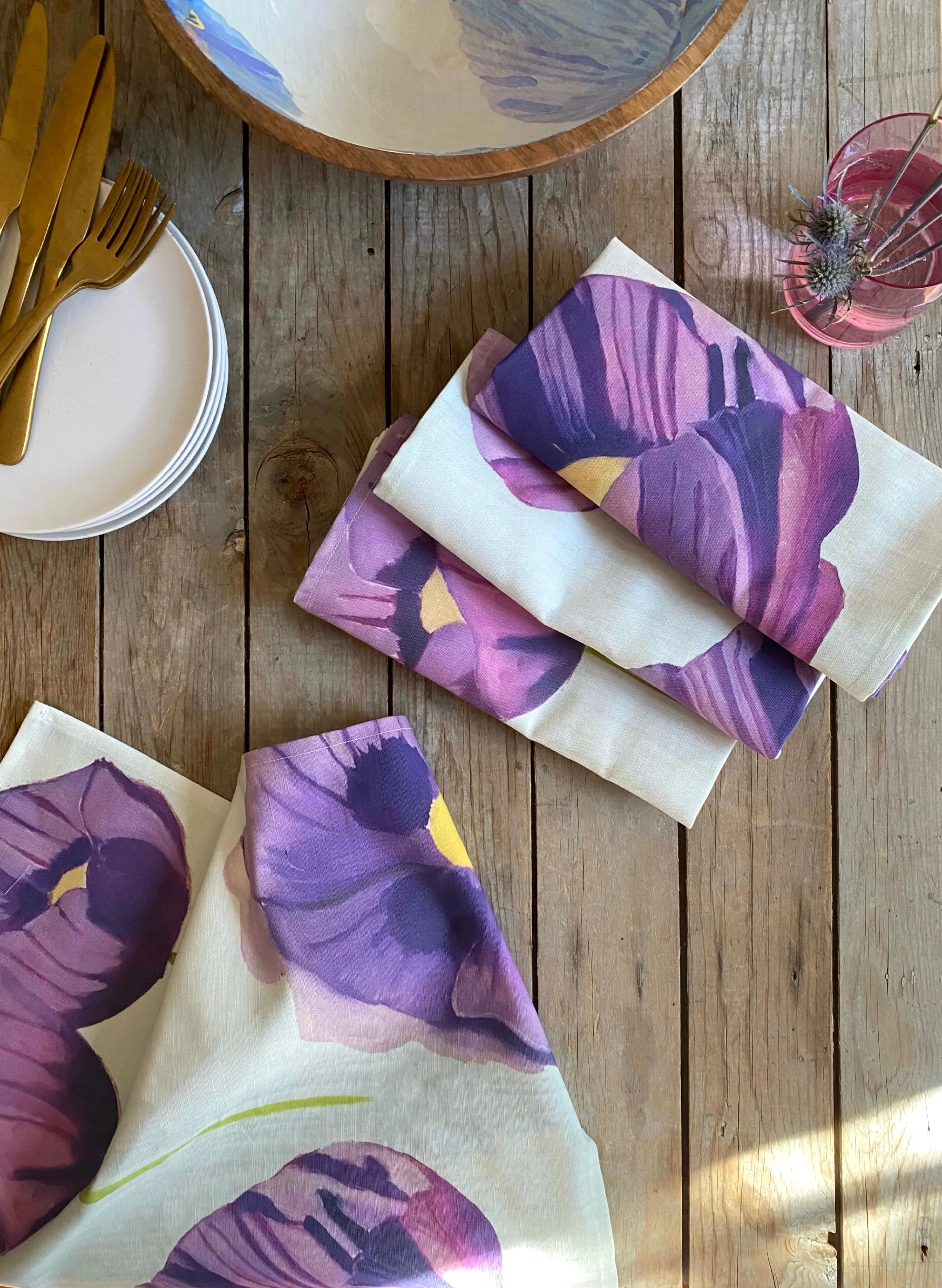 Rustic wooden table with folded floral purple and white table linens, gold cutlery, white plates, and a vase with dried flowers