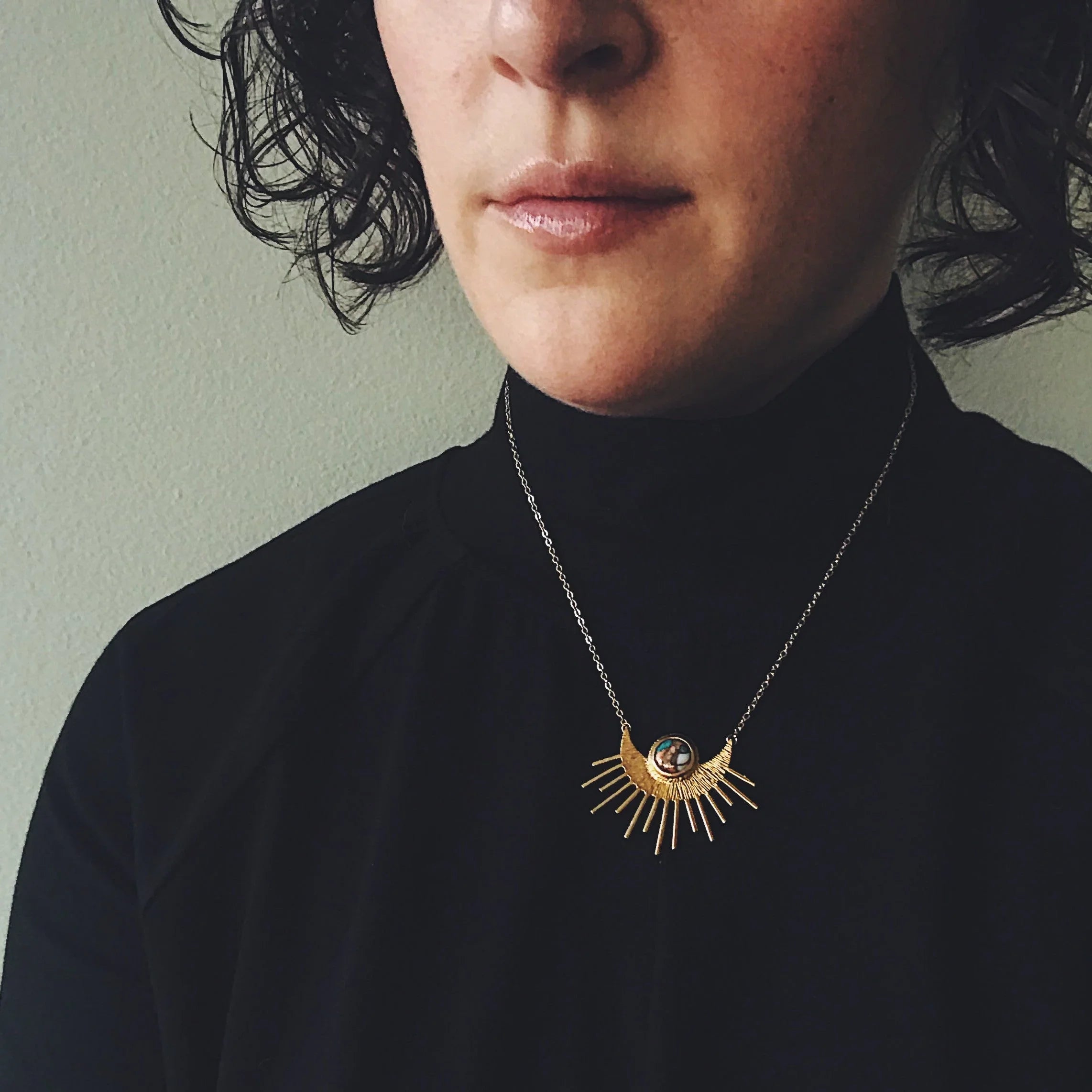 Woman wearing a black turtleneck and a gold sunburst pendant necklace with a central gemstone
