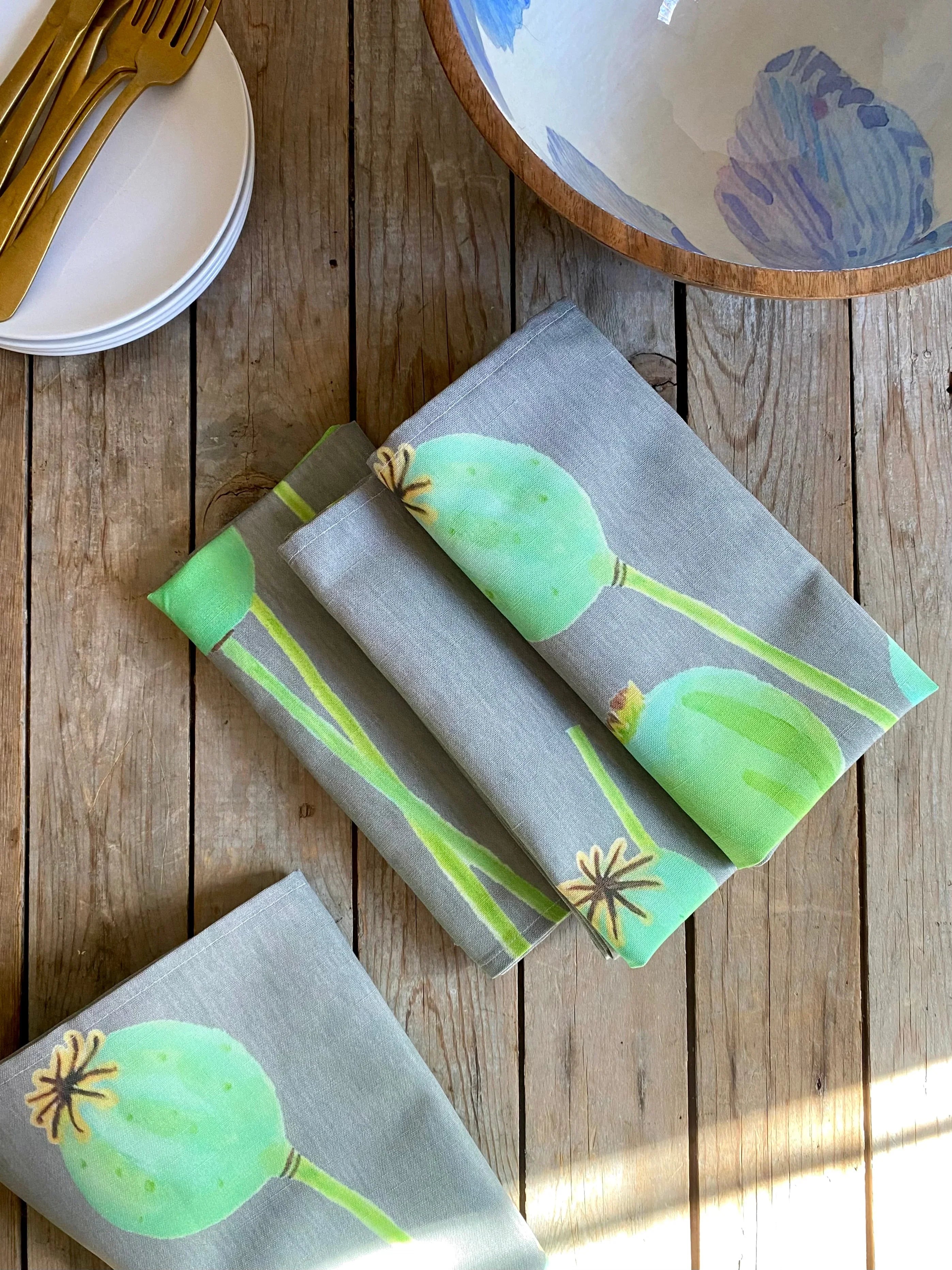 Folded gray cloth napkins with green botanical print on rustic wooden table, white plates, gold utensils