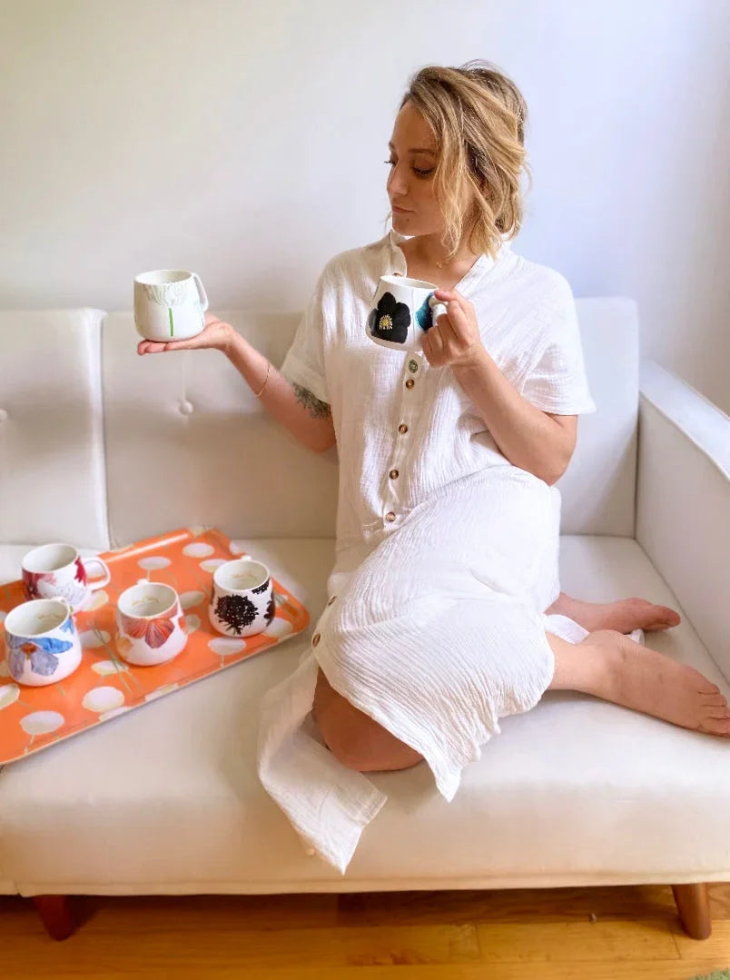 Woman in white dress sitting on white couch holding decorative floral mugs with colorful mug tray beside her